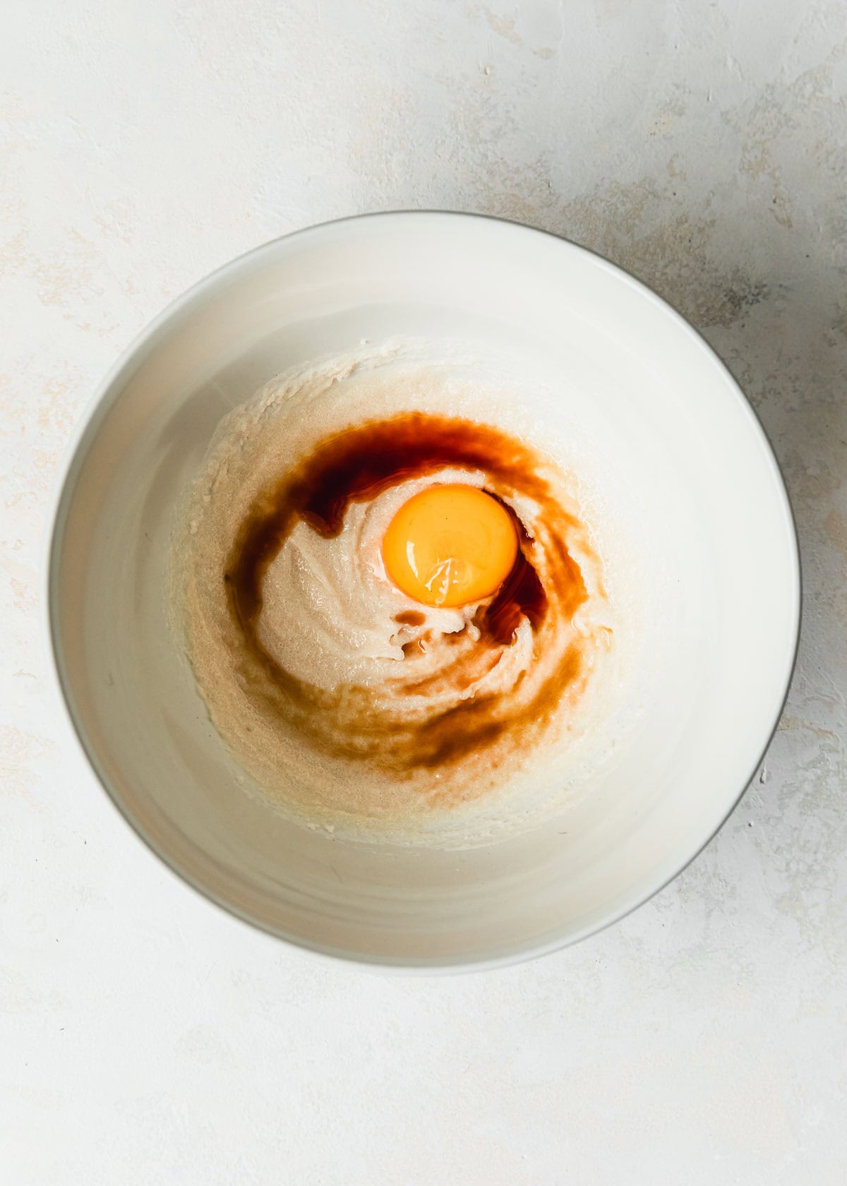 A white bowl of butter, sugar, an egg yolk, and vanilla on a white stone counter.