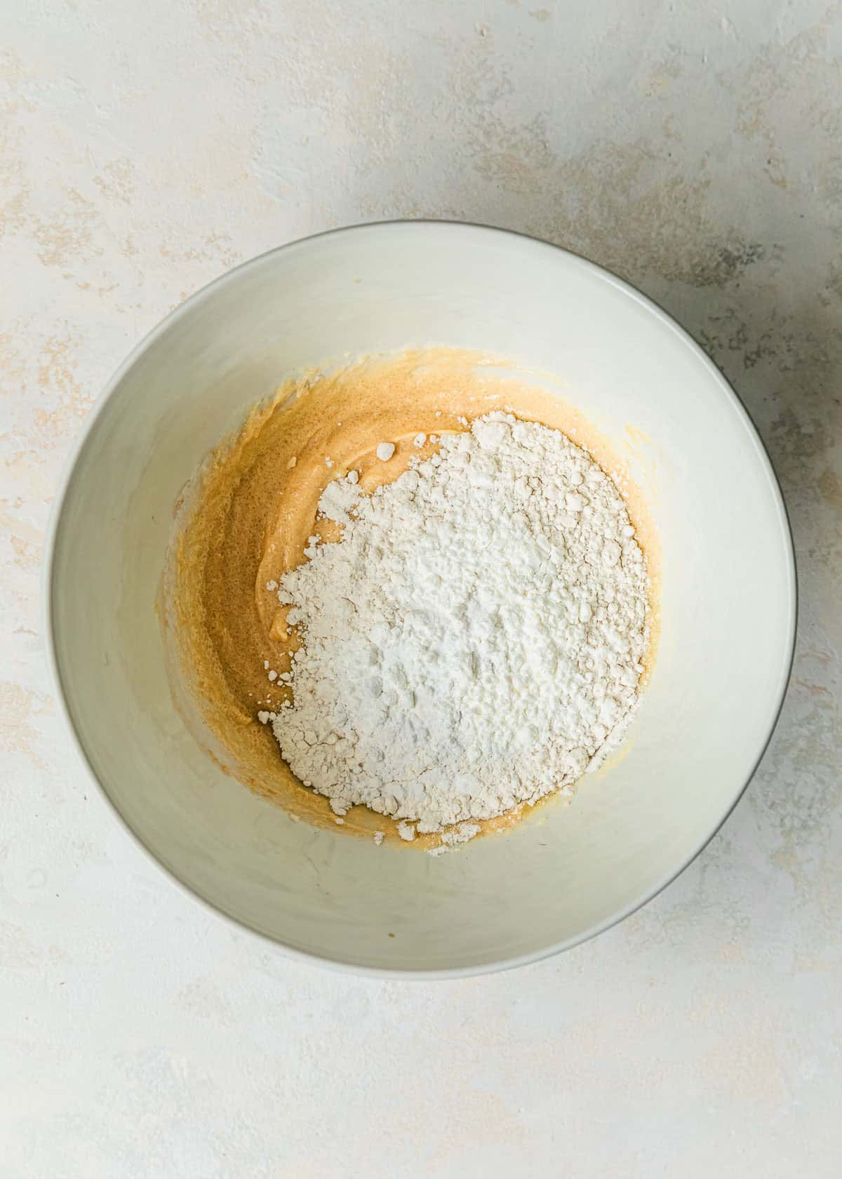 A white bowl of sugar, butter, an egg yolk, vanilla, and flour on a white table.