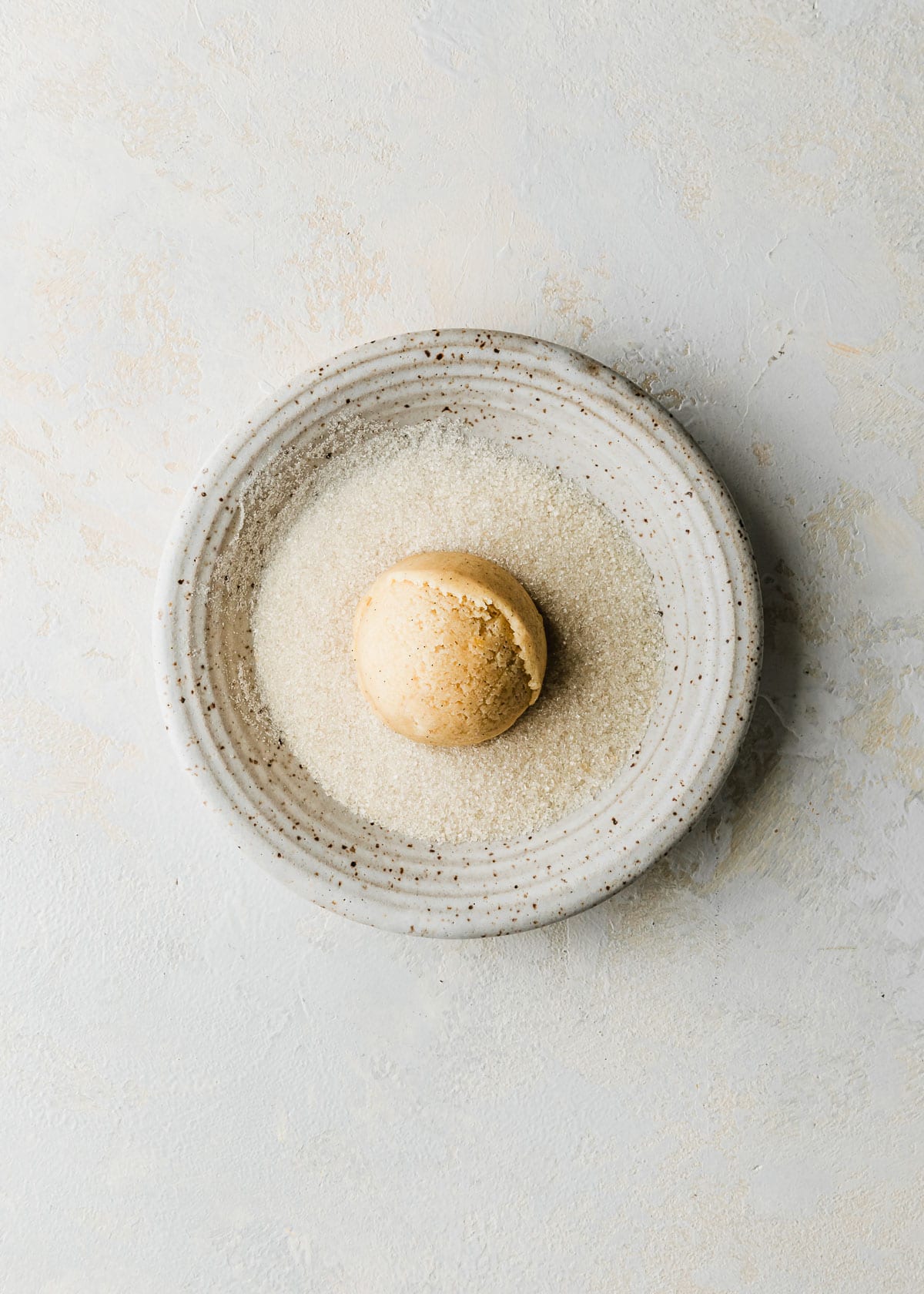 A white bowl of sugar with a ball of cookie dough on a white counter.