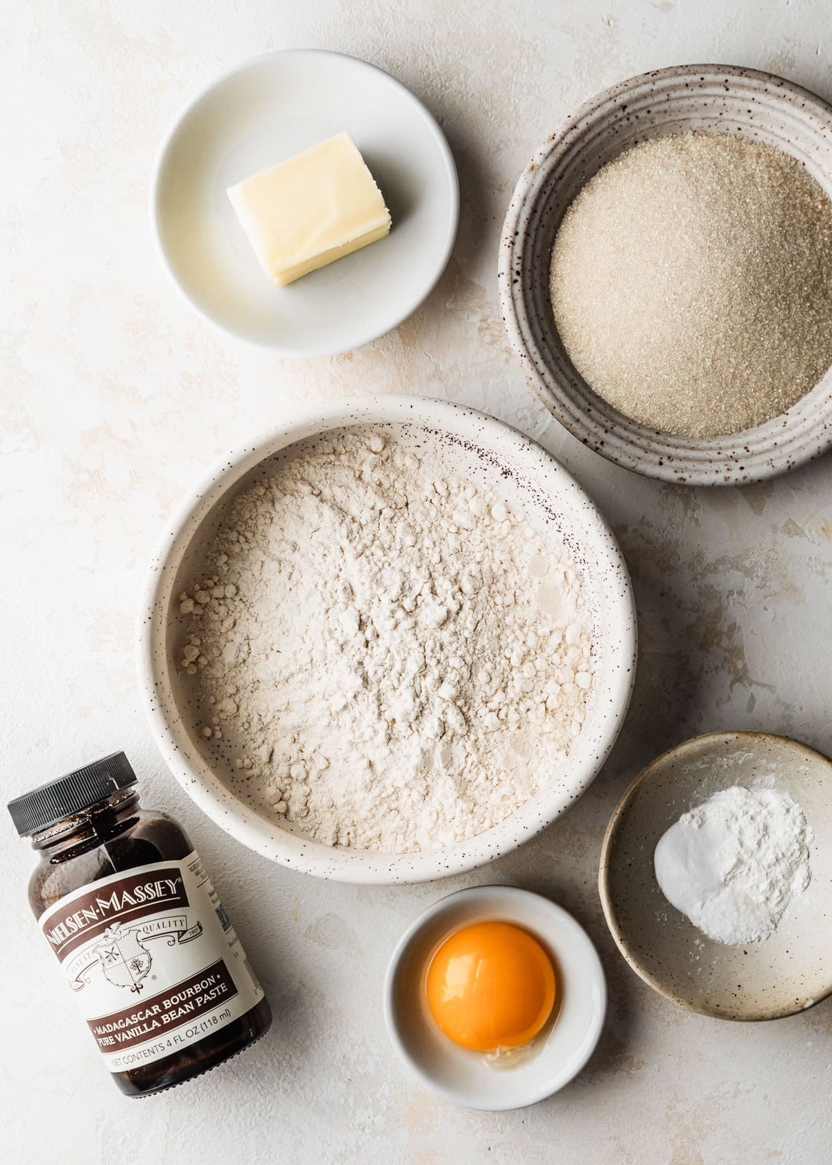 White bowls of flour, sugar, an egg yolk, cornstarch, and butter on a tan counter next to a jar of vanilla bean paste.