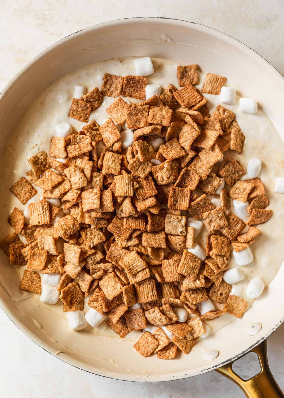 A white skillet of melted marshmallows and Cinnamon Toast Crunch on a tan background.
