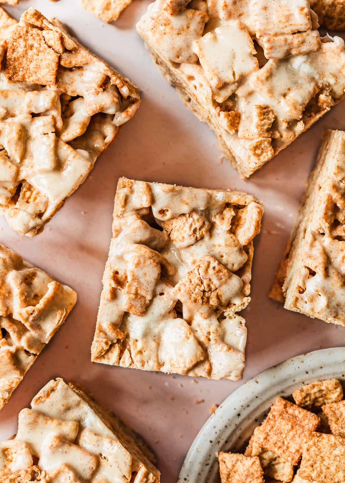 Rows of small batch cinnamon toast crunch marshmallow treats next to a white bowl of cereal with a pink background.