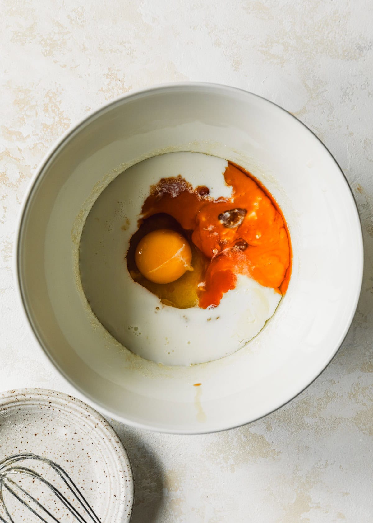 A white bowl of buttermilk, butter, sugar, an egg, and vanilla on a beige counter.