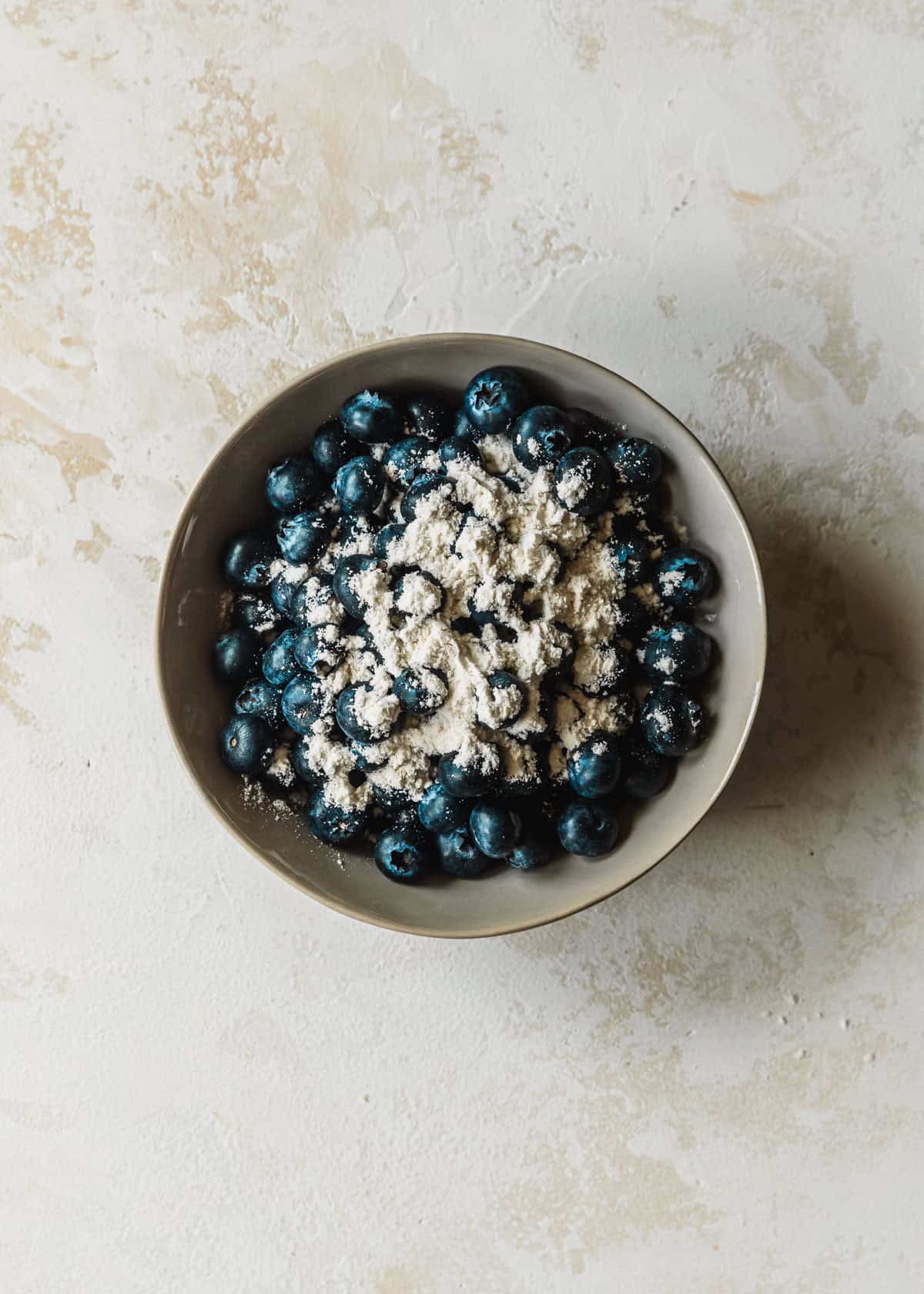A grey bowl of blueberries and flour on a tan counter.