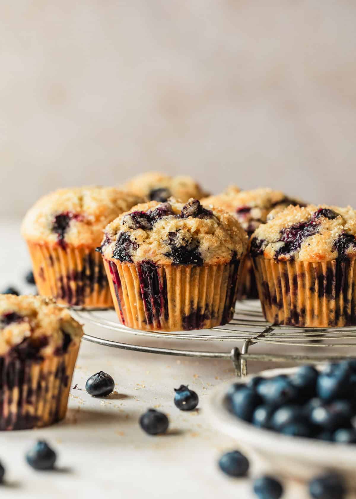 A wire rack of small batch blueberry muffins next to a white bowl of berries with a tan background.