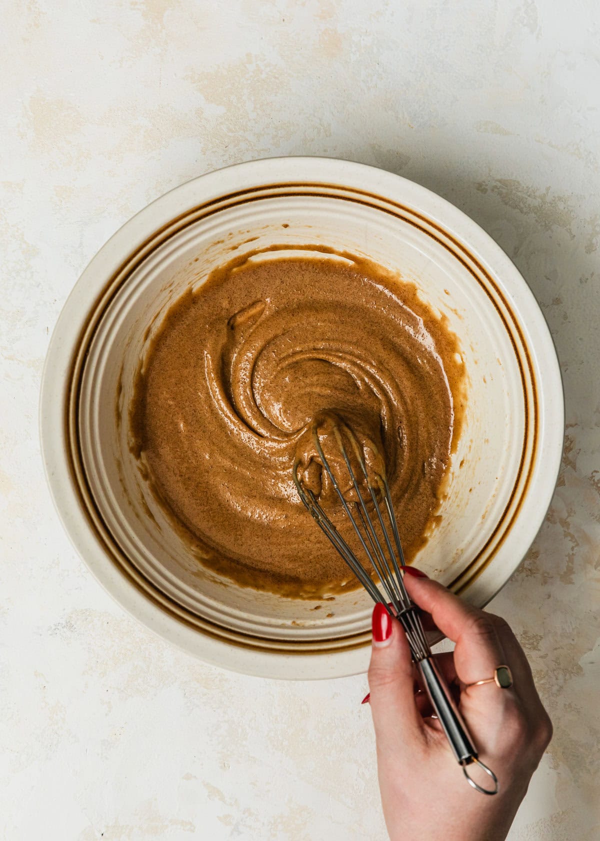A hand using a whisk to stir a white bowl of brown butter, brown sugar, and an egg yolk.