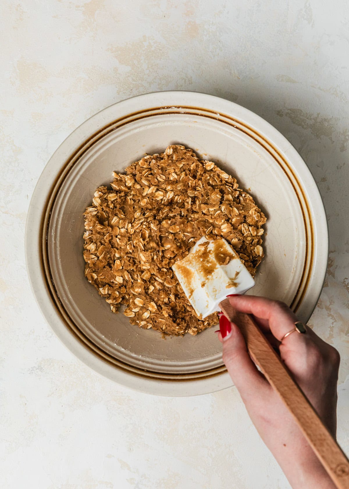 A hand using a rubber spatula to stir a white bowl of dough for small batch oatmeal cookies.