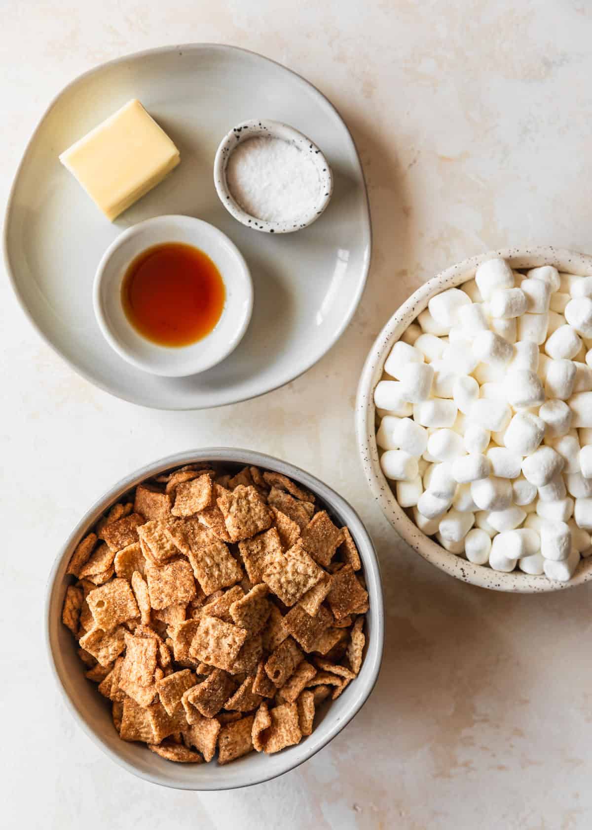 White bowls of vanilla, butter, salt, marshmallows, and cinnamon cereal on a tan counter.