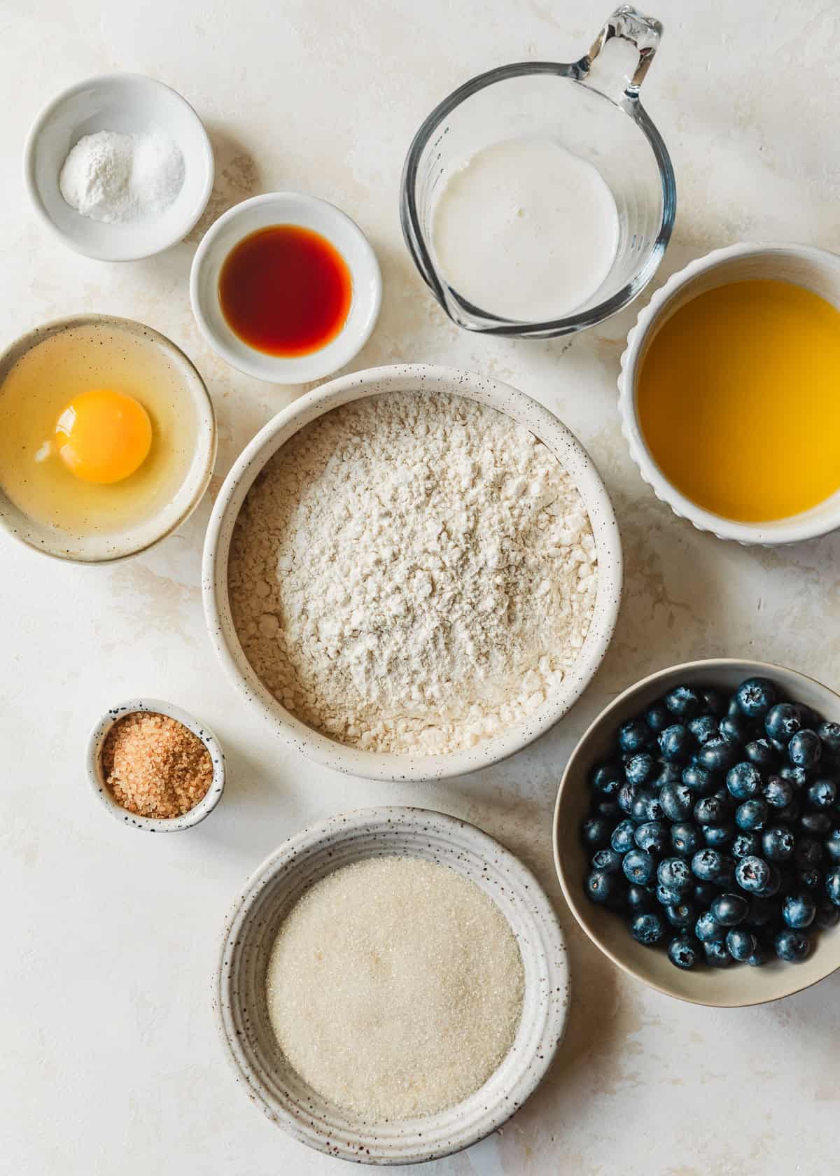 White bowls of flour, butter, vanilla, an egg, baking powder, sugar, and blueberries on a tan counter.