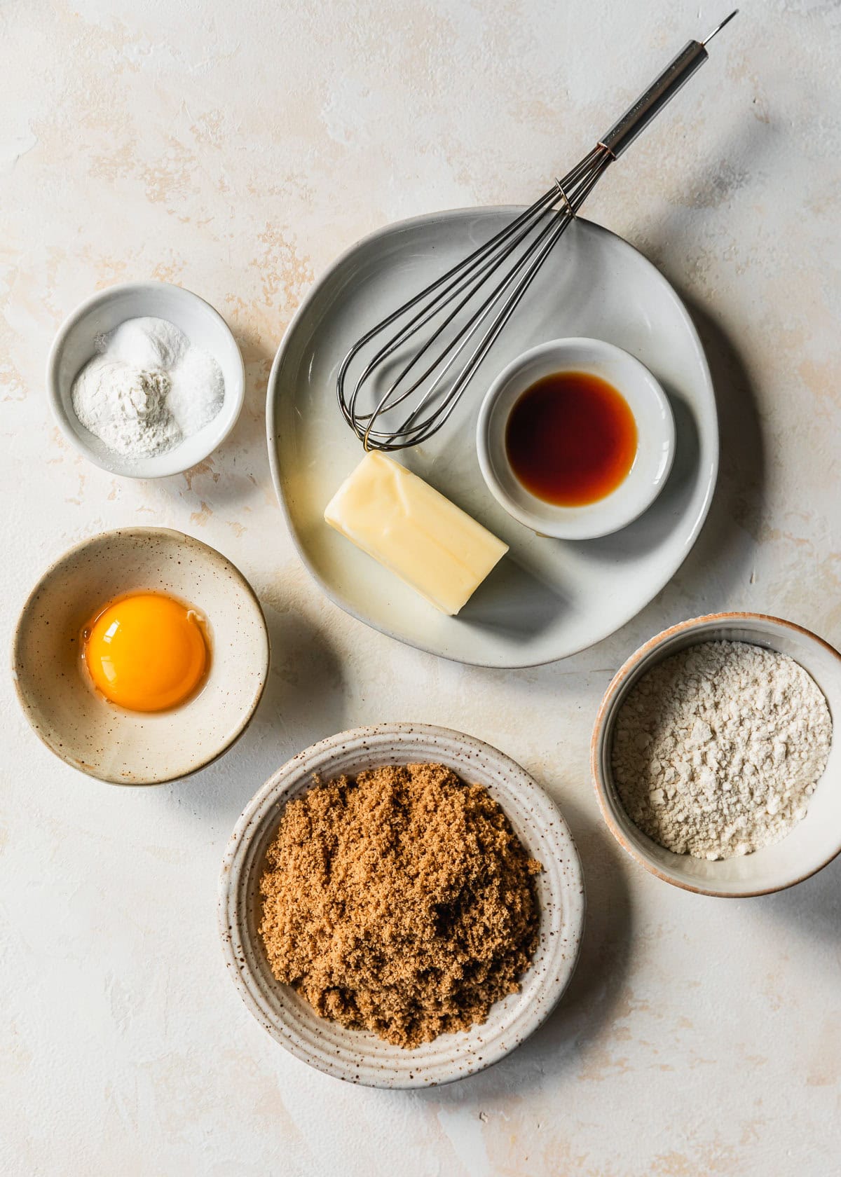 White bowls of butter, vanilla, cornstarch, baking soda, salt, an egg yolk, brown sugar, and flour on a tan counter.