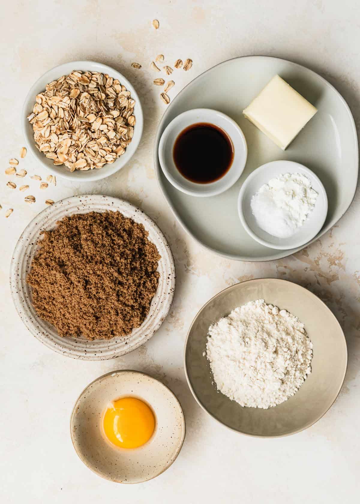 White bowls of oats, vanilla, butter, cornstarch, salt, baking soda, brown sugar, flour, and an egg yolk on a tan counter.