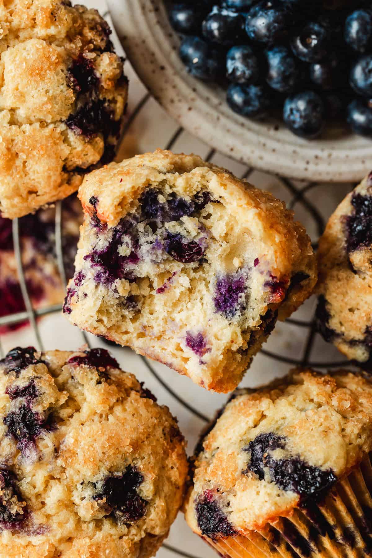 A wire rack of small batch blueberry muffins and a white bowl of blueberries on a tan counter.