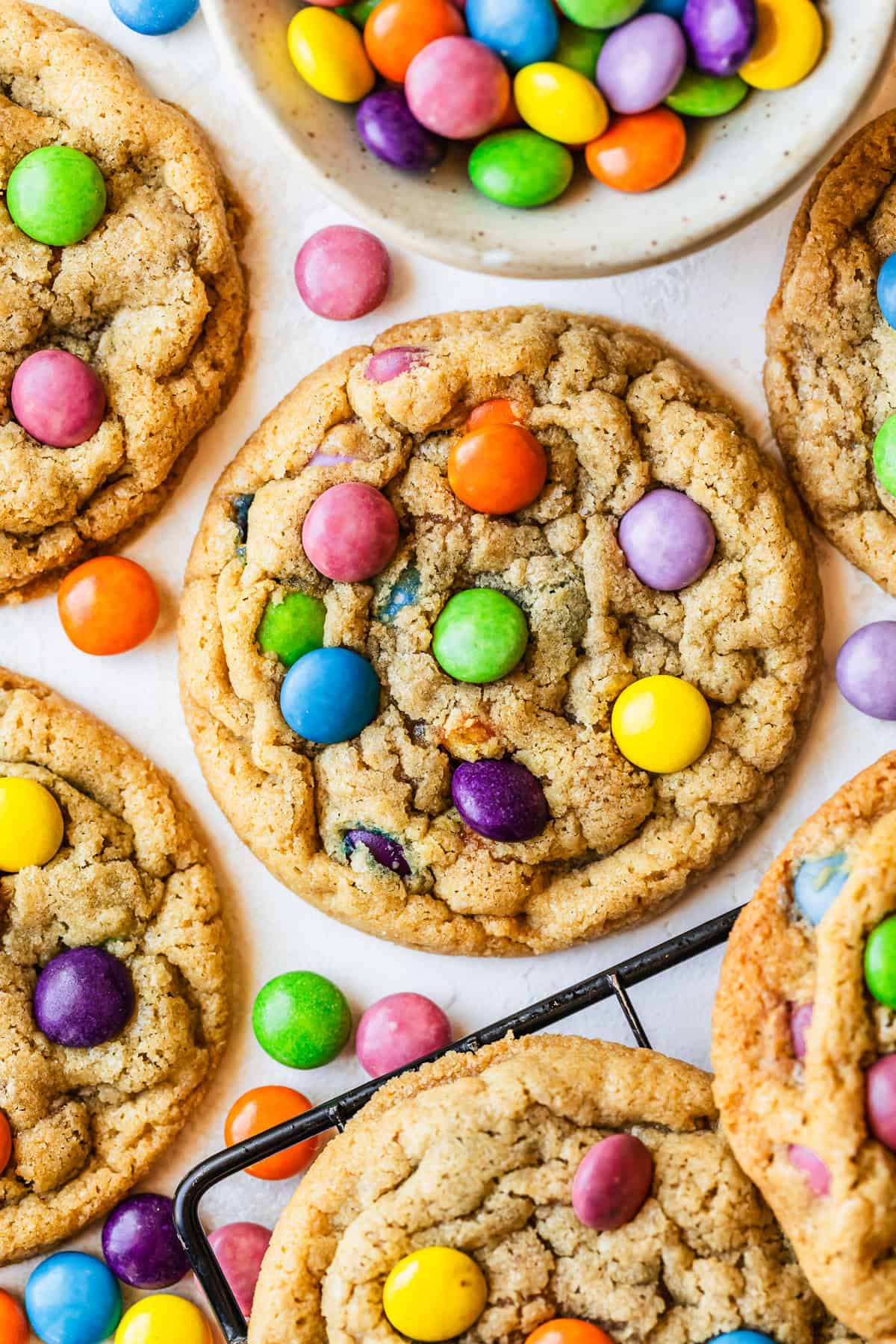Rows of small batch M&M cookies next to a wire rack of cookies and a white bowl of M&Ms on a tan counter.