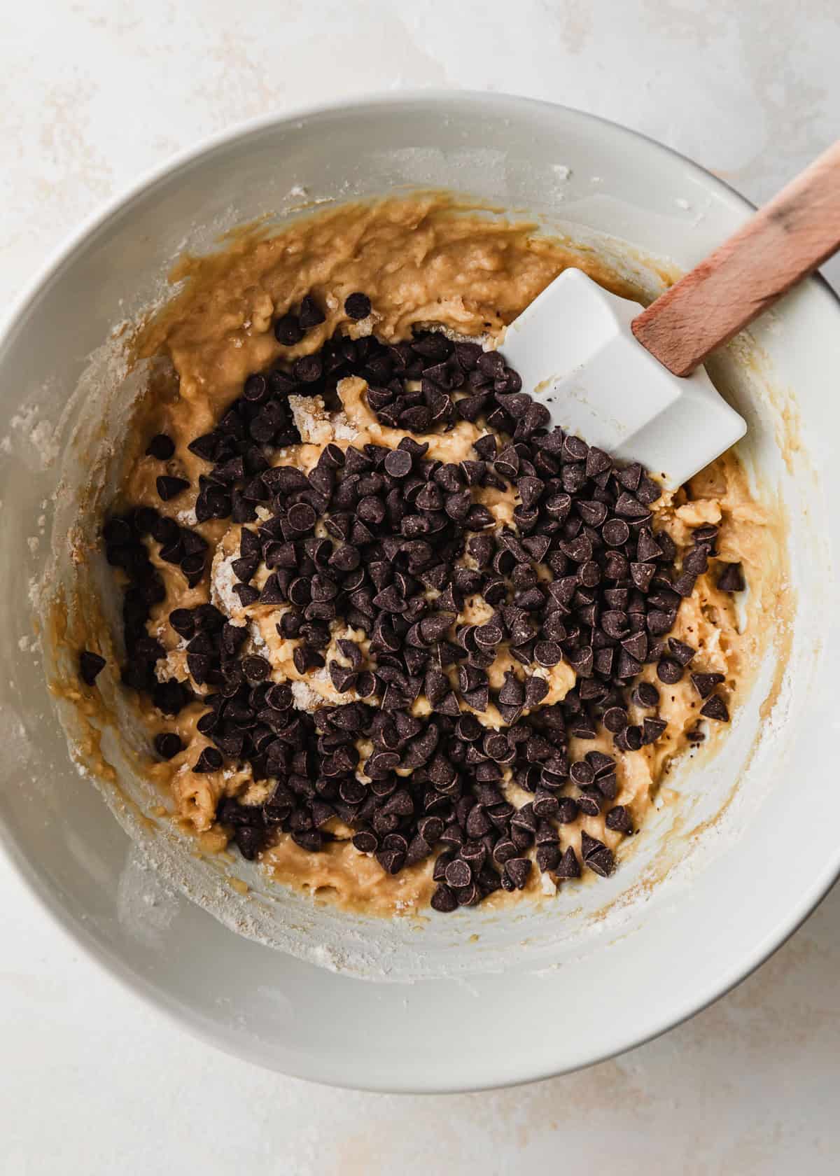 A rubber spatula mixing mini chocolate chips into a white bowl of small batch chocolate chip muffins batter.