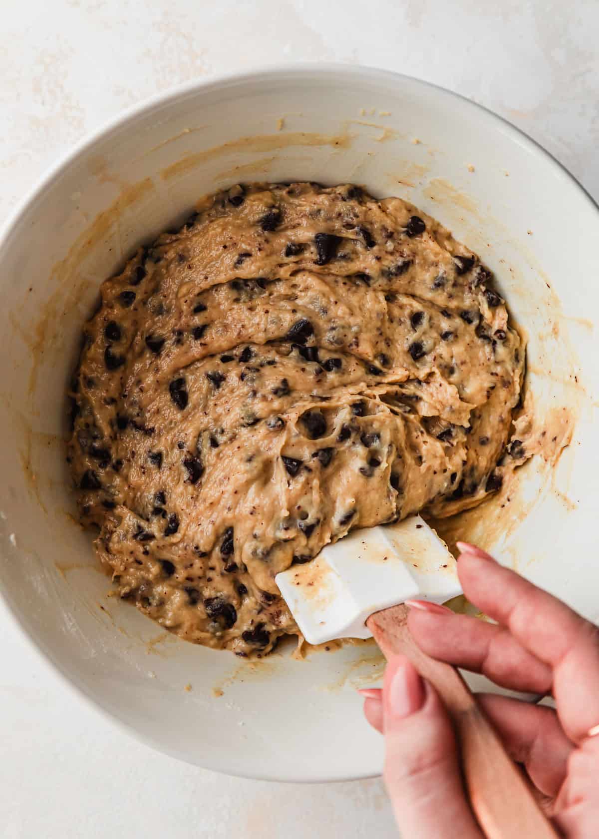 A hand using a rubber spatula to mix batter for small batch chocolate chip muffins in a white bowl.
