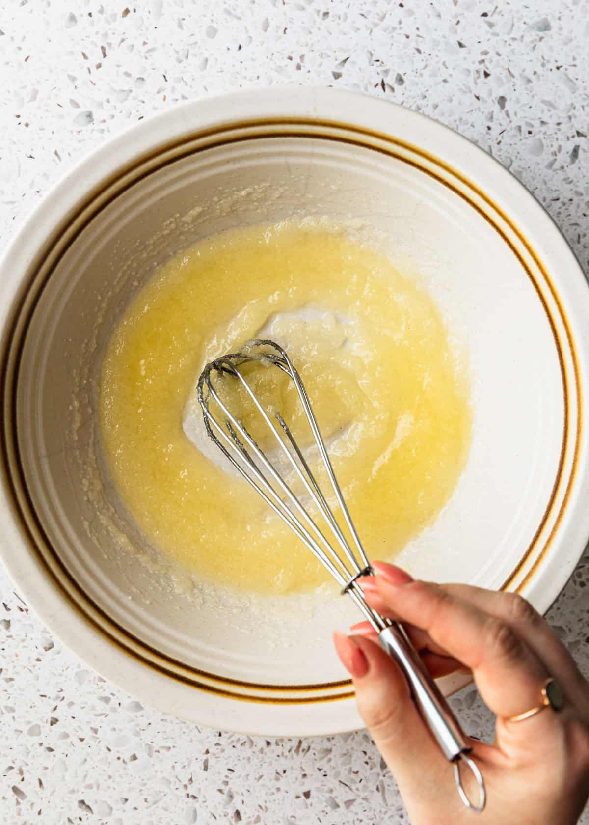 A hand using a whisk to mix butter and sugar in a white bowl.