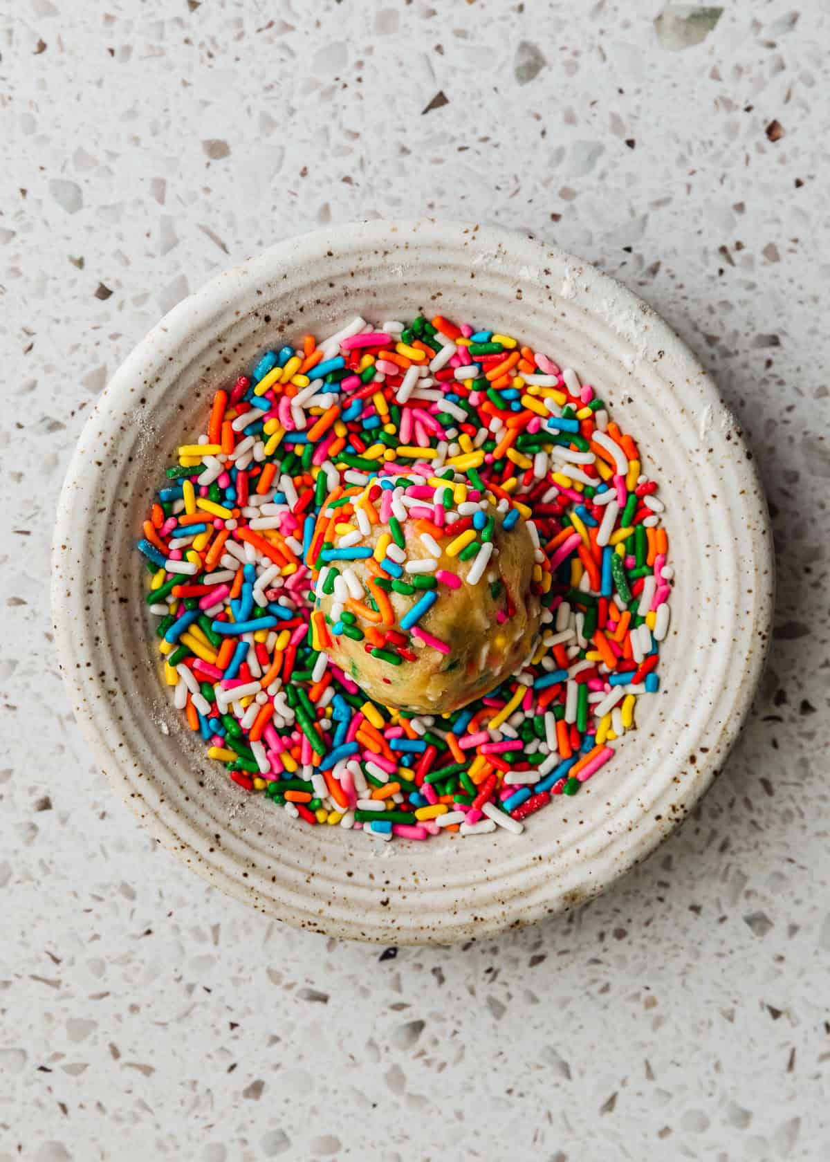 A white bowl of sprinkles with a scoop of cookie dough on a white terrazzo counter.