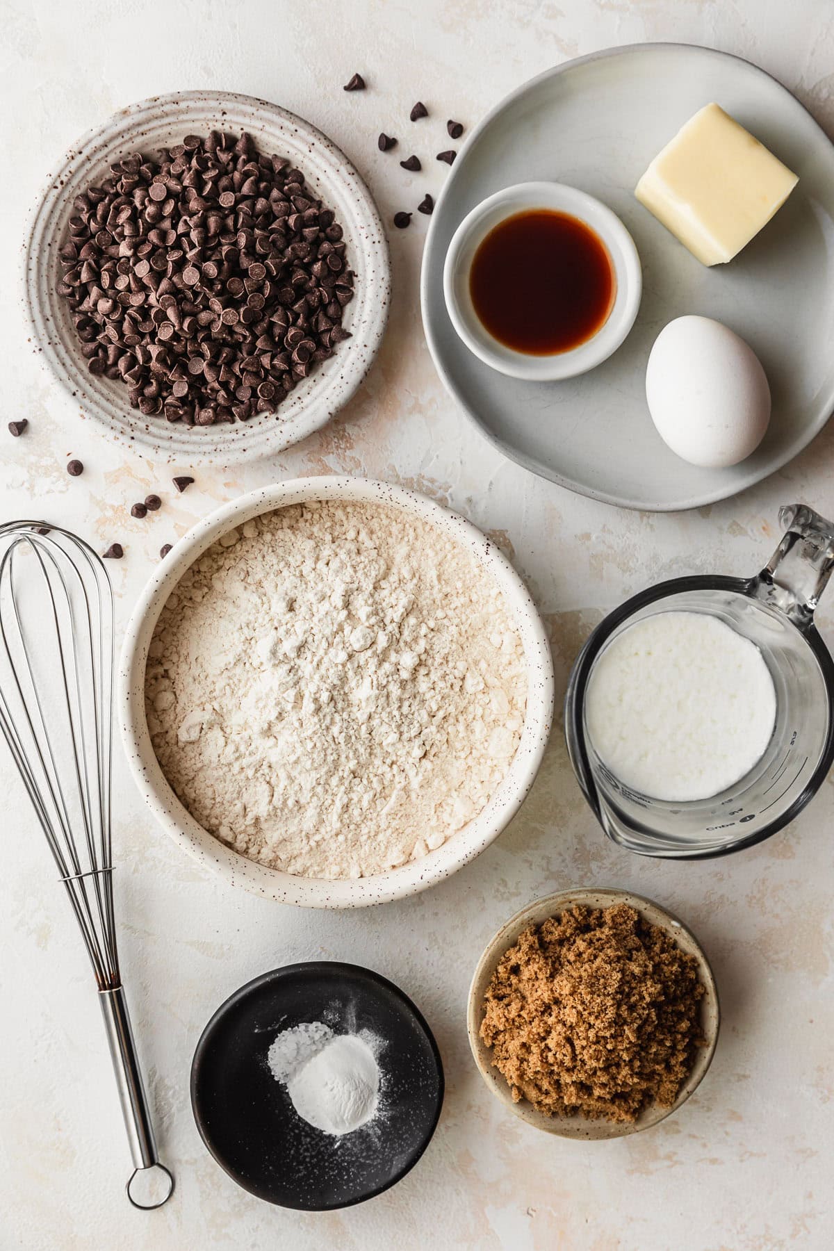 White and black bowls of flour, brown sugar, baking powder, salt, buttermilk, vanilla, an egg, and mini chocolate chips on a tan counter.