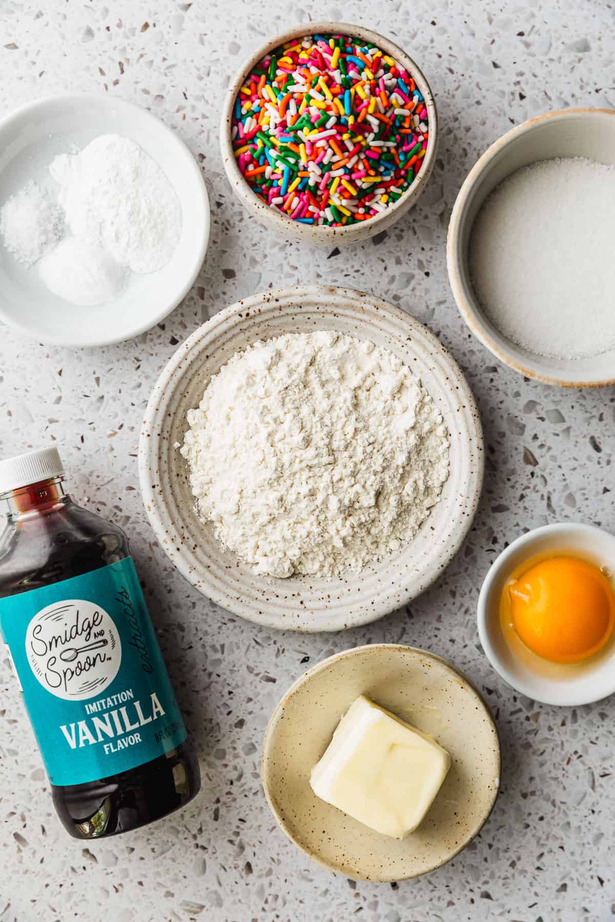 White bowls of flour, sugar, an egg yolk, butter, rainbow sprinkles, baking soda, salt, and cornstarch on a white counter next to a bottle of artificial vanilla.