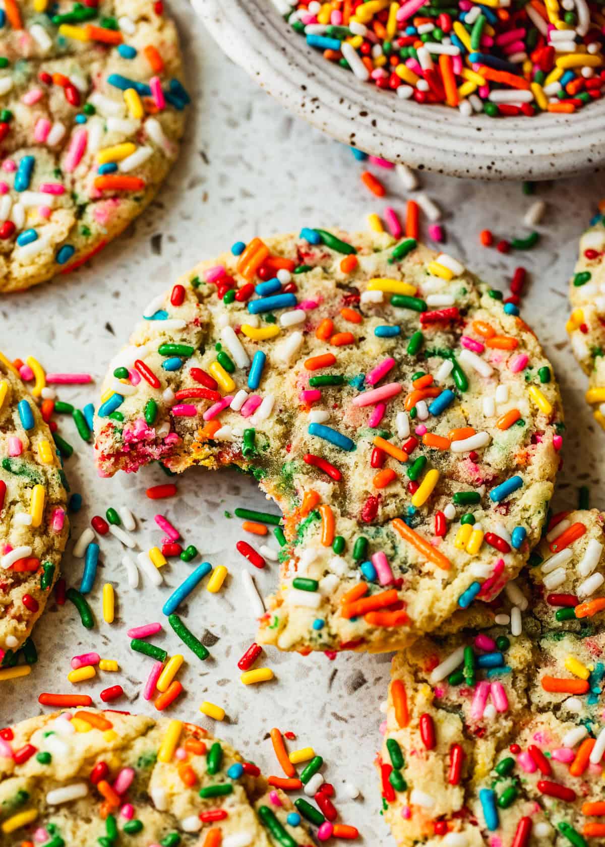 A cookie with a bite taken out of it next to rows of small batch funfetti cookies and a white bowl of rainbow sprinkles on a white terrazzo counter.