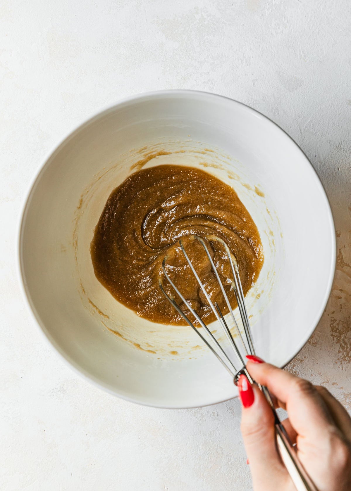 A hand using a whisk to mix butter and sugars in a white bowl.