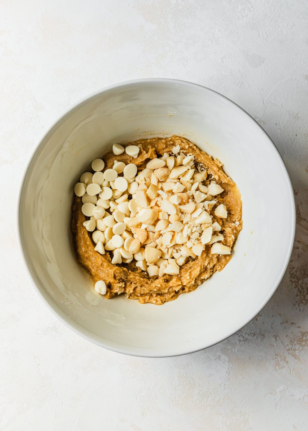 A white bowl of cookie dough with white chocolate chips and macadamia nuts.