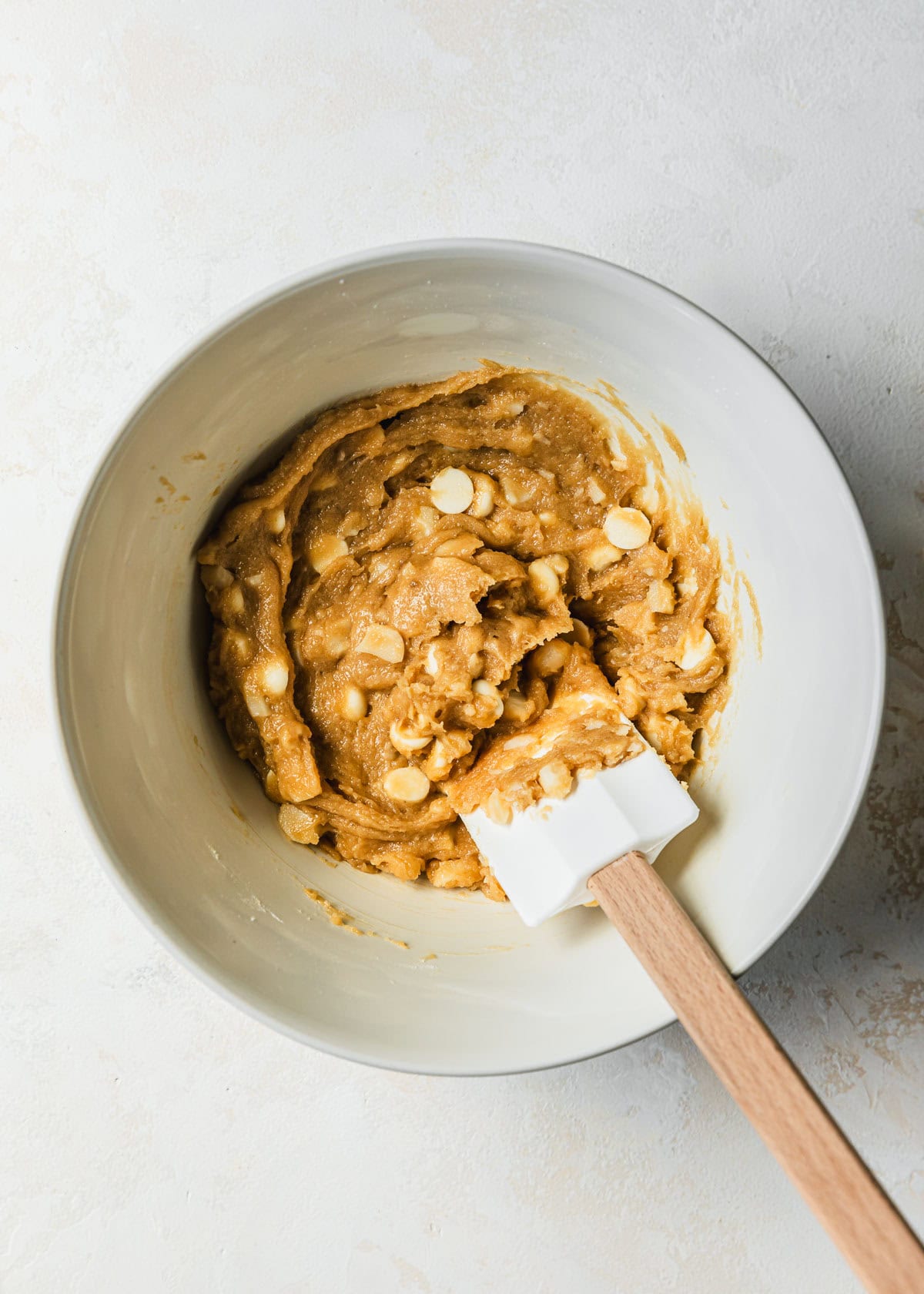 A rubber spatula mixing dough for small batch white chocolate macadamia nuts in a white bowl.