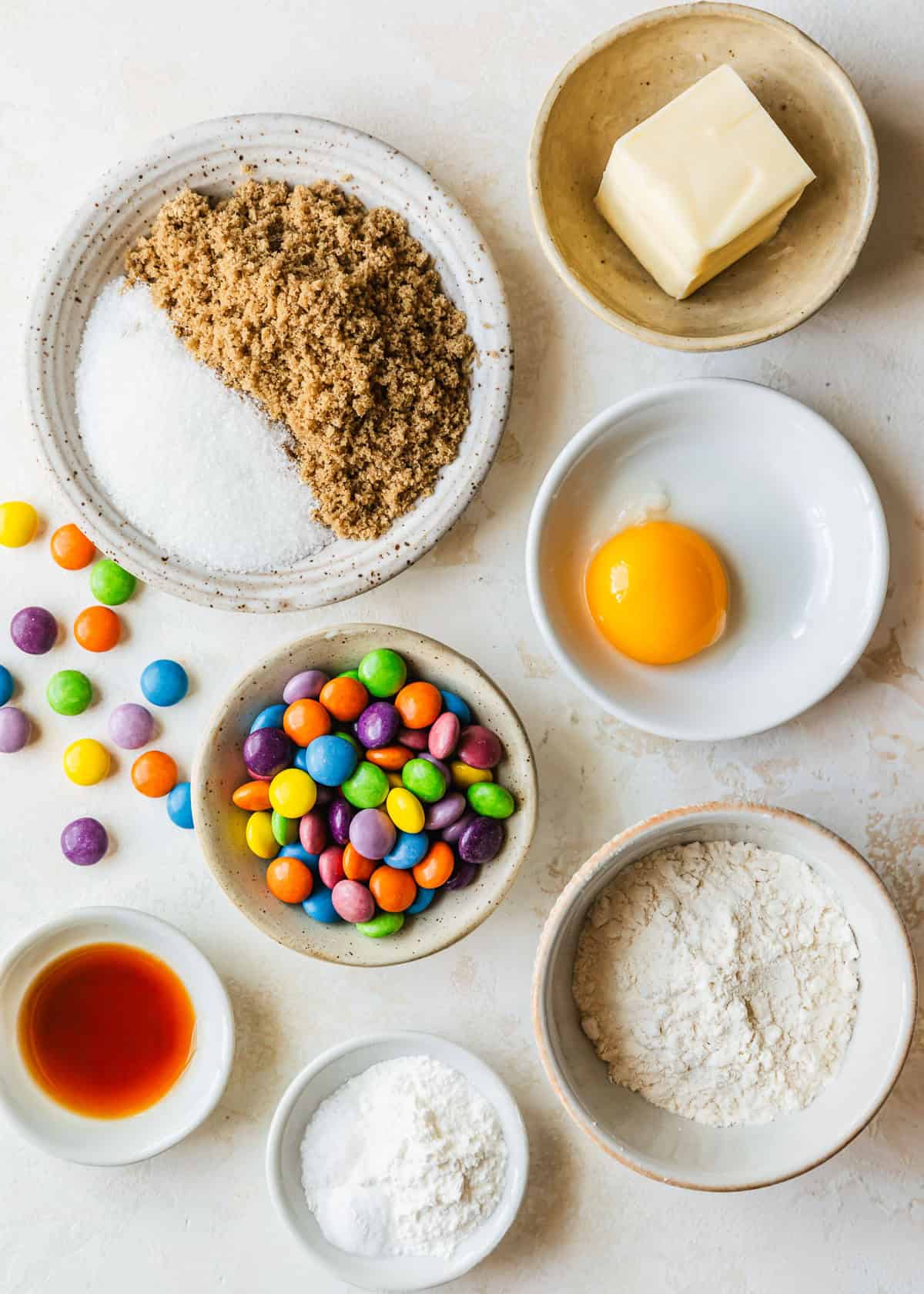 White bowls of sugar, butter, an egg yolk, flour, cornstarch, baking soda, salt, vanilla, and rainbow candies on a tan counter.