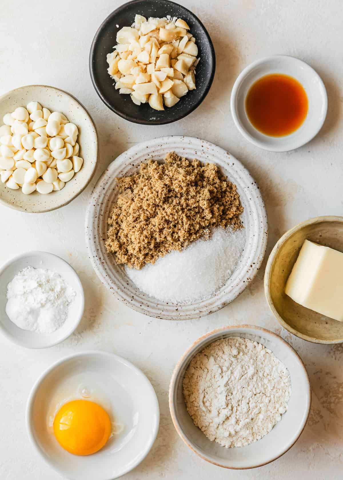 White and black bowls of nuts, vanilla chips, vanilla, brown and white sugar, cornstarch, salt, baking soda, butter, flour, and an egg on a tan counter.