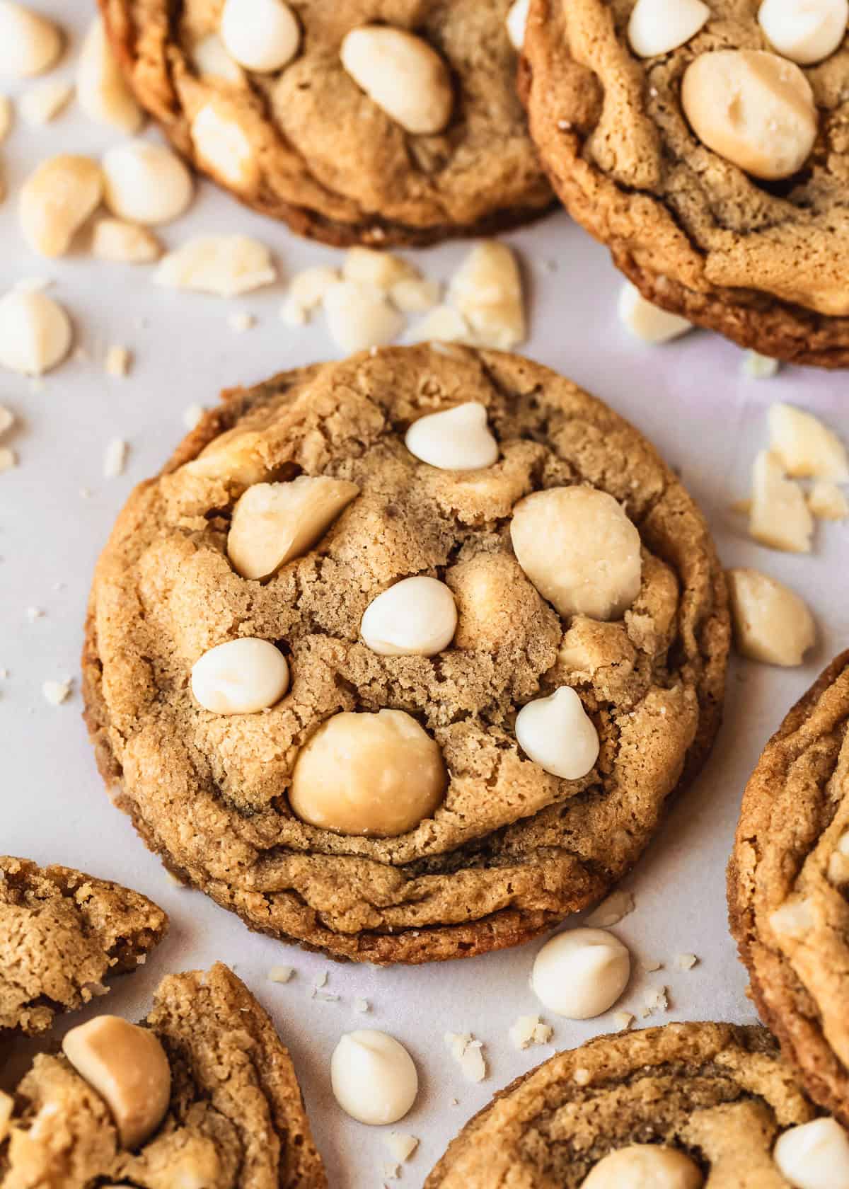 A small batch of white chocolate macadamia nut cookies on a pink counter next to white chocolate chips & nuts.
