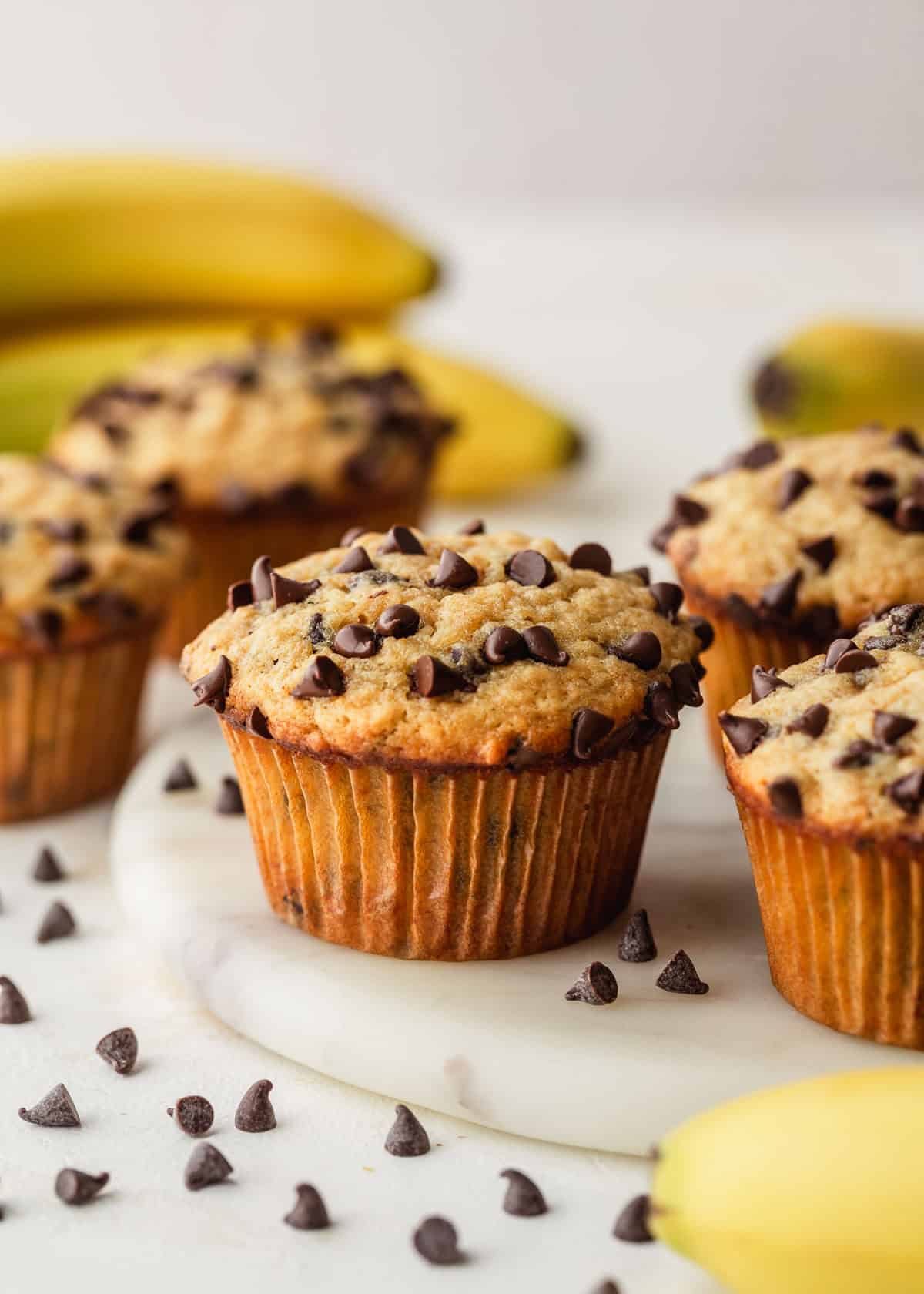 Rows of small batch chocolate chip banana muffins on a white platter next to bananas on a white backdrop.