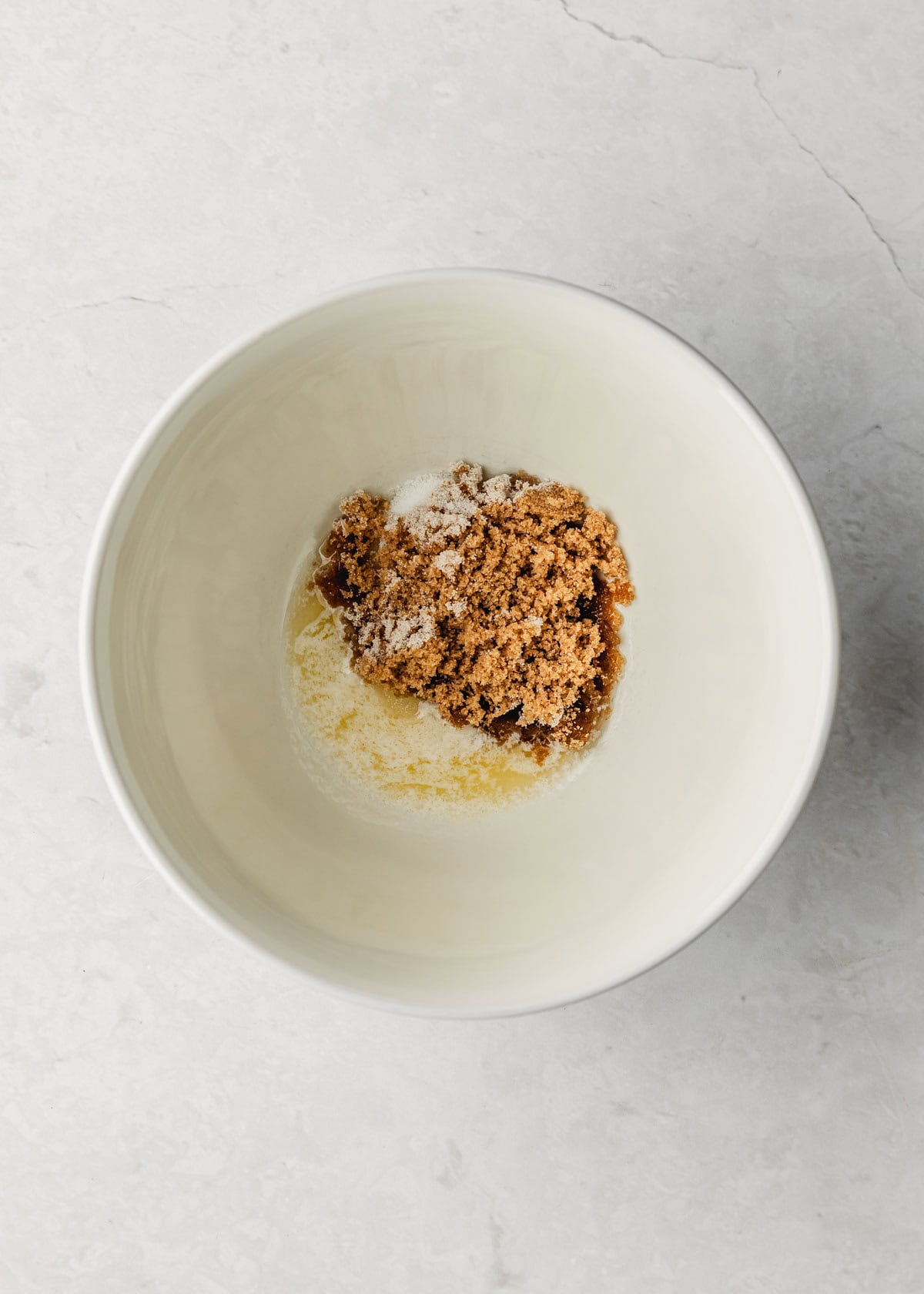 A white bowl of melted butter, brown sugar, and sugar on a white counter.