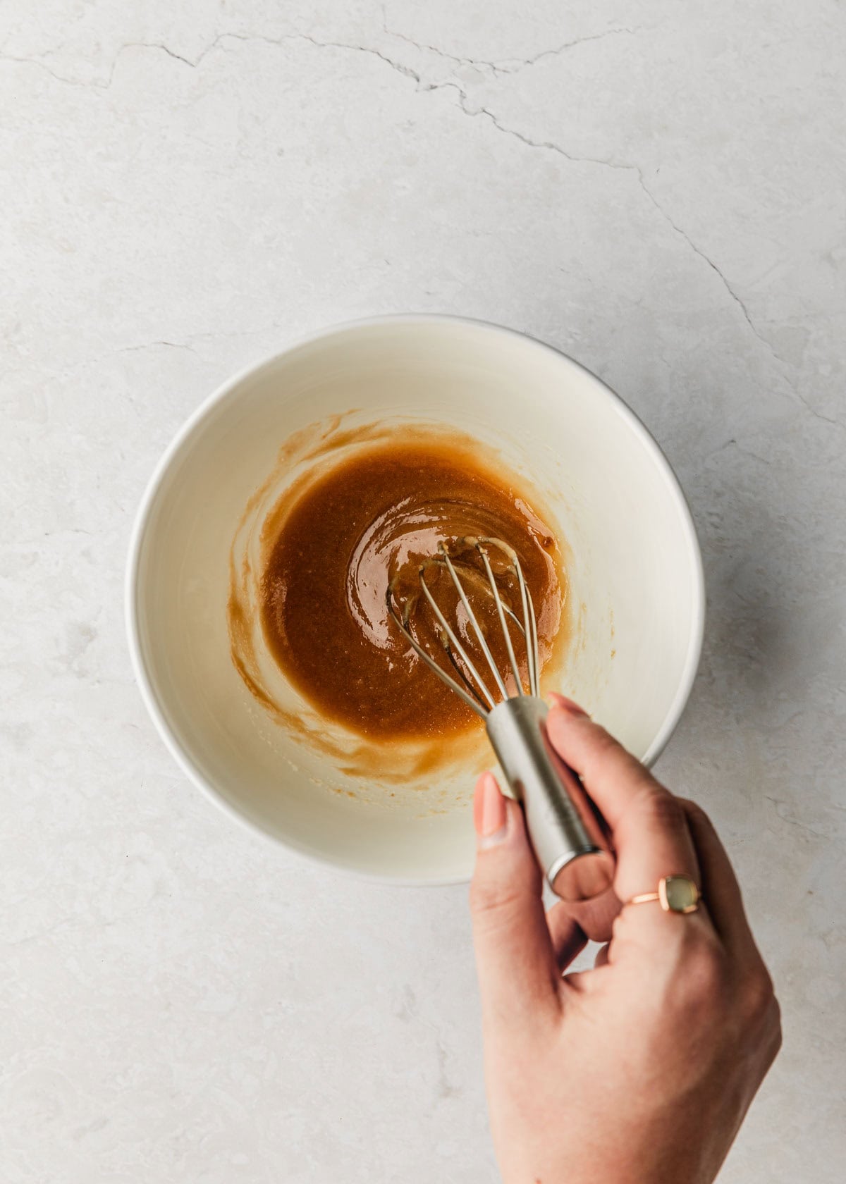 A hand using a whisk to mix butter, sugar, and cream in a white bowl on a white counter.