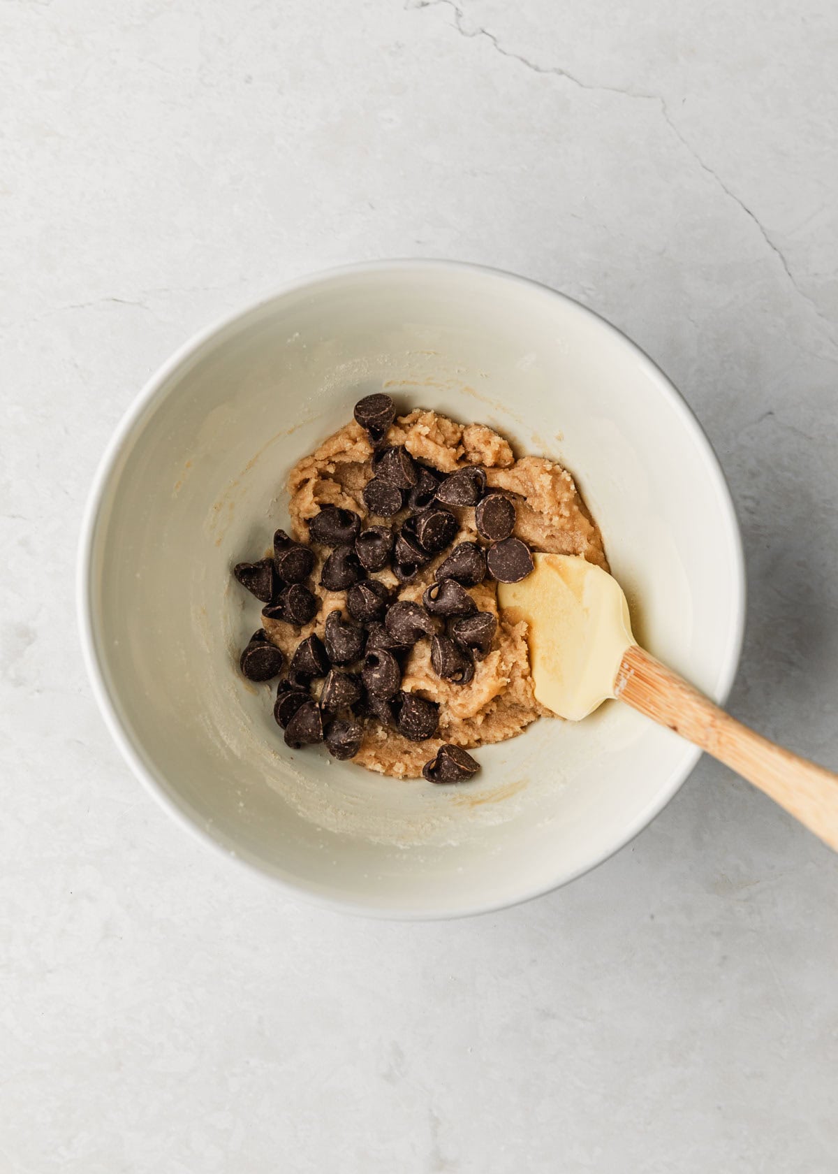 A white bowl of dough for one chocolate chip cookie on a white table.