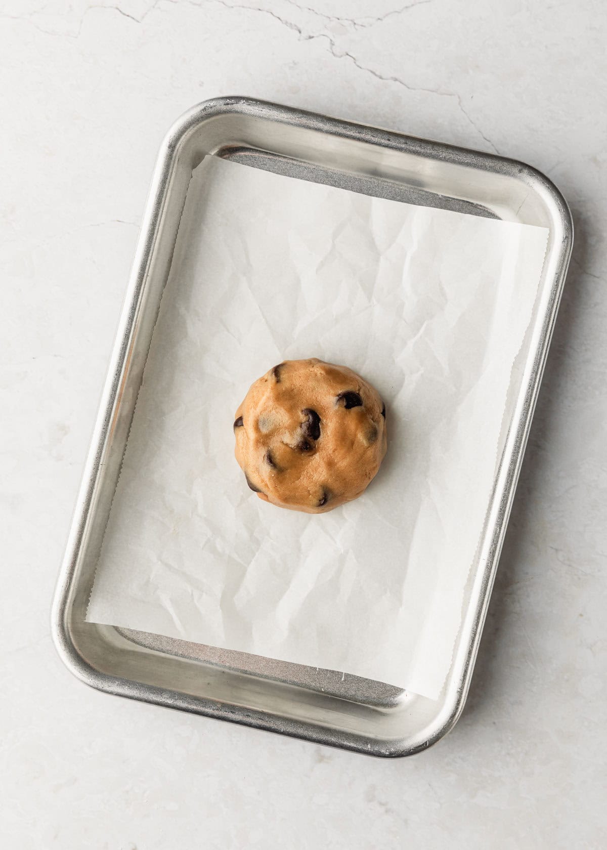 A sheet pan with a raw single serving chocolate chip cookie on a marble table.