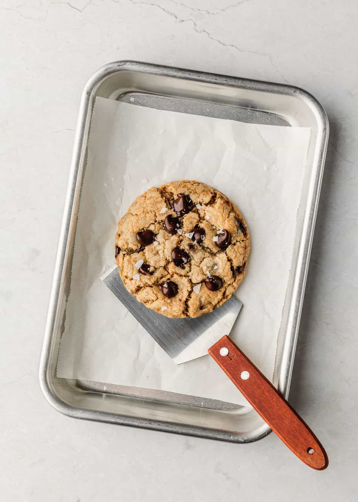 A sheet pan with one chocolate chip cookie and a spatula on a marble counter.