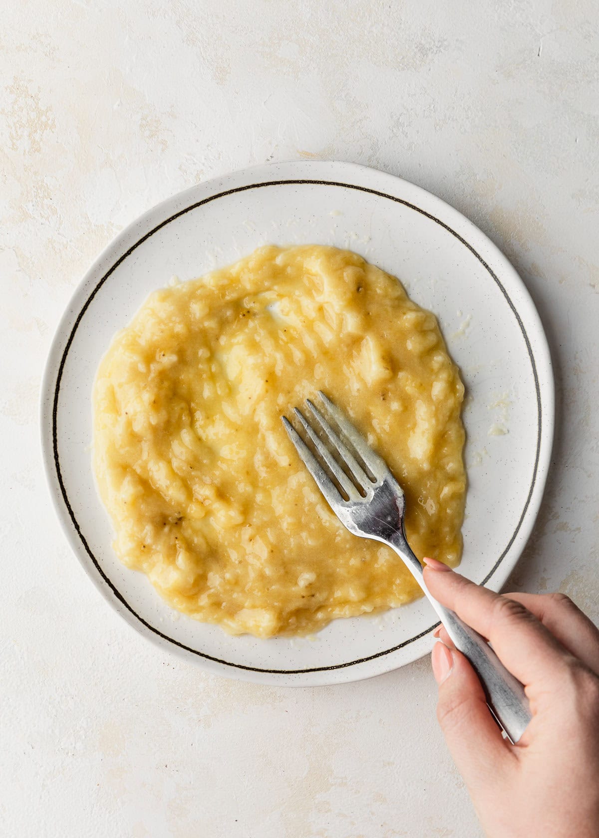 A hand using a fork to mash a banana on a white plate on a white background.