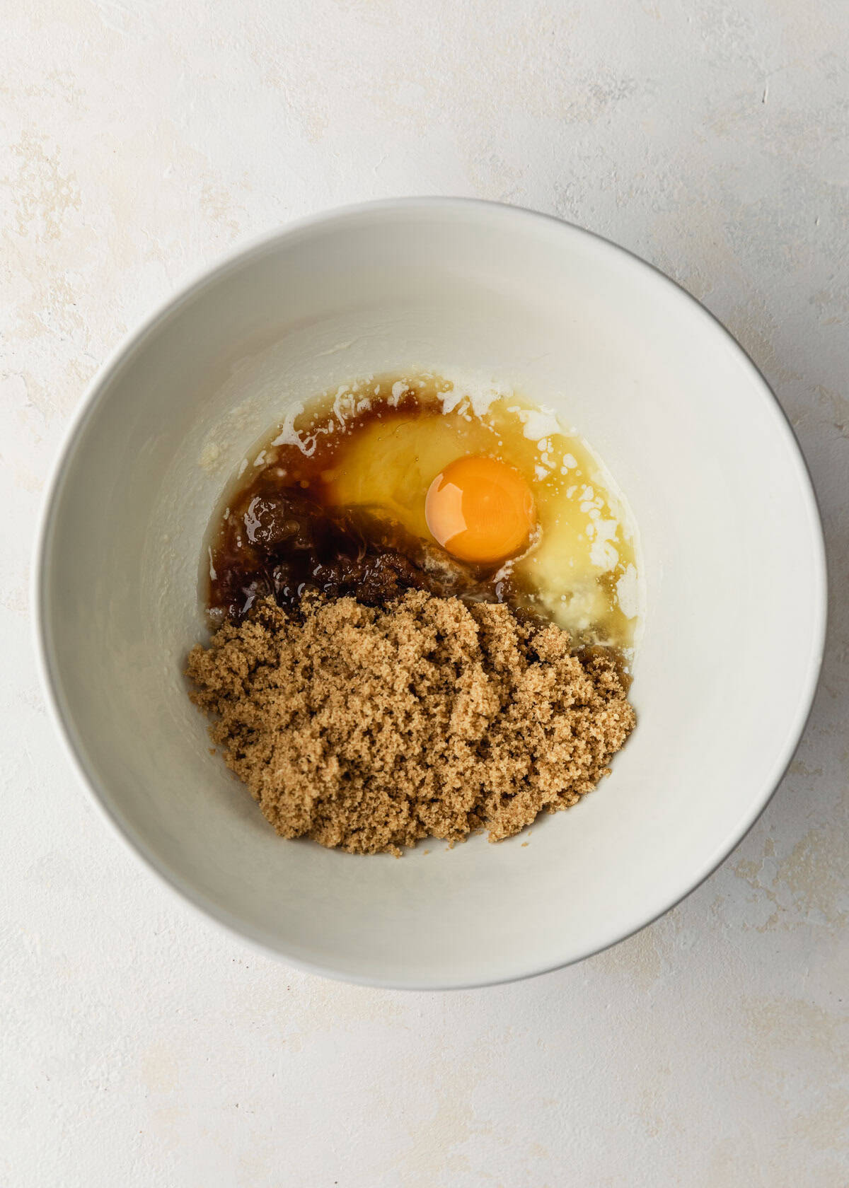 A white bowl of butter, sugar, vanilla, and an egg on a white counter.