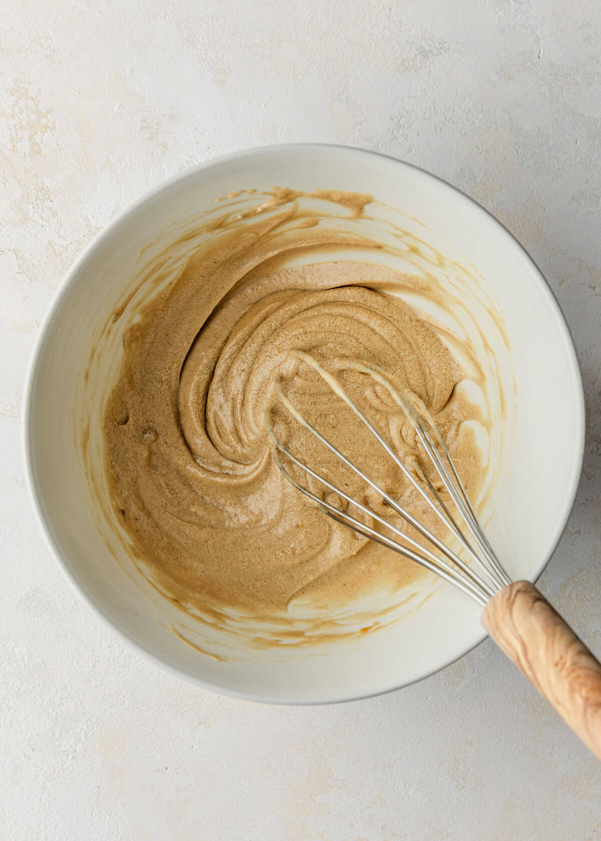 A white bowl of whisked butter and sugar and a whisk on a white counter.