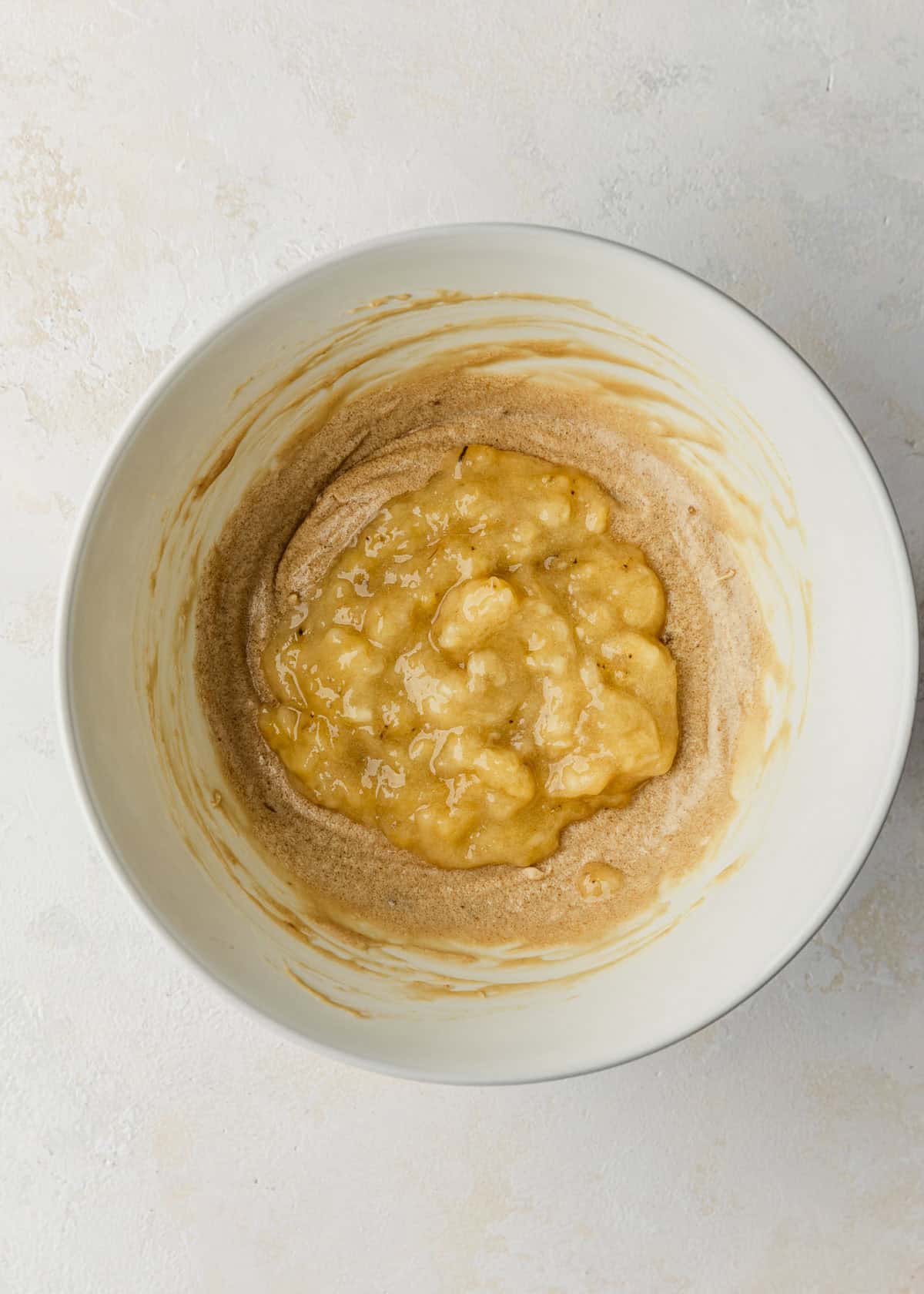 A white bowl of whisked butter, sugar, and mashed banana on a white table.