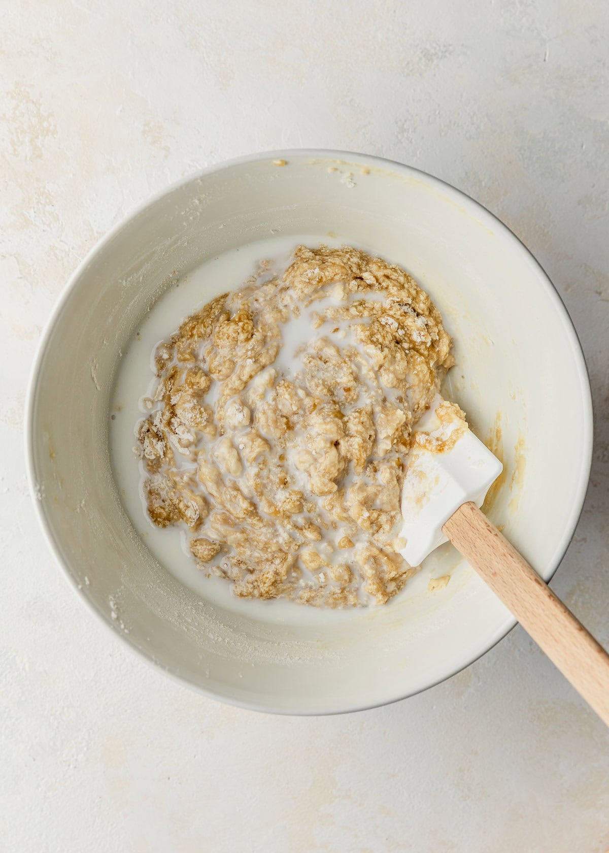 A white bowl of batter for small batch chocolate chip banana muffins and milk on a white counter.