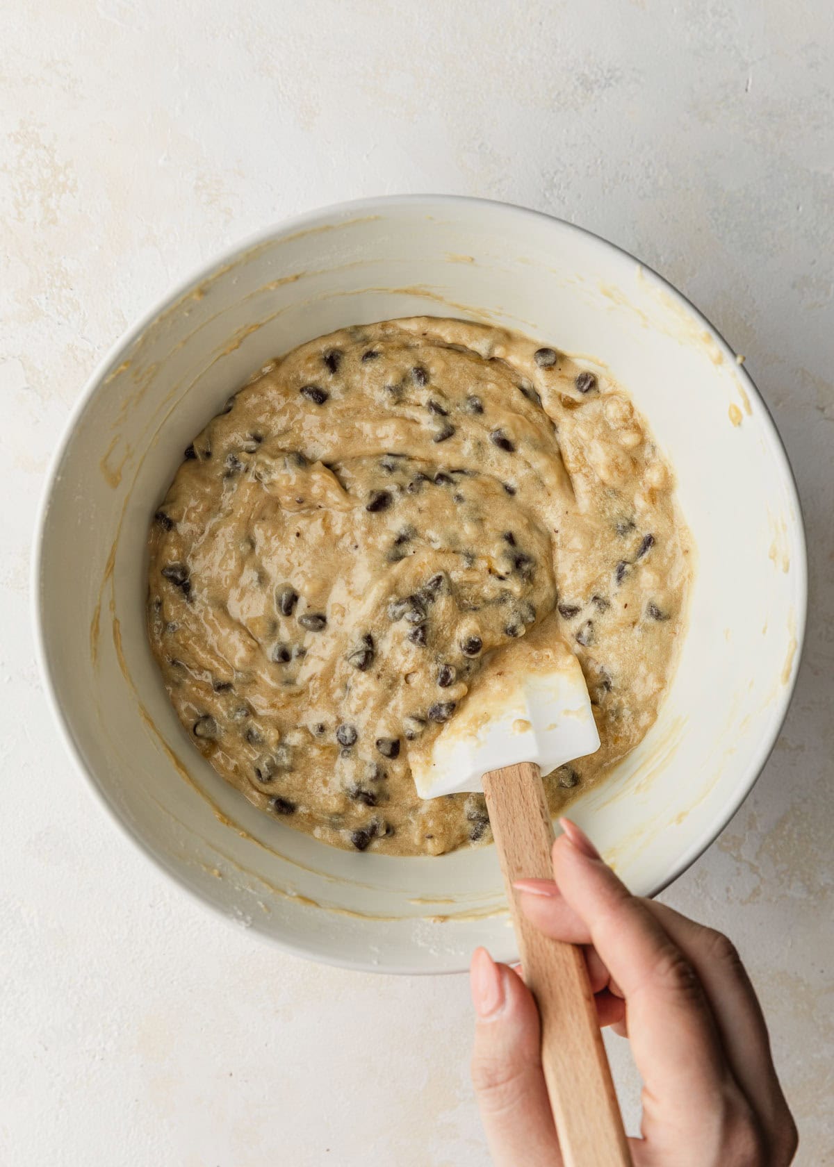 A hand using a rubber spatula to mix batter for small batch chocolate chip banana muffins in a white bowl.