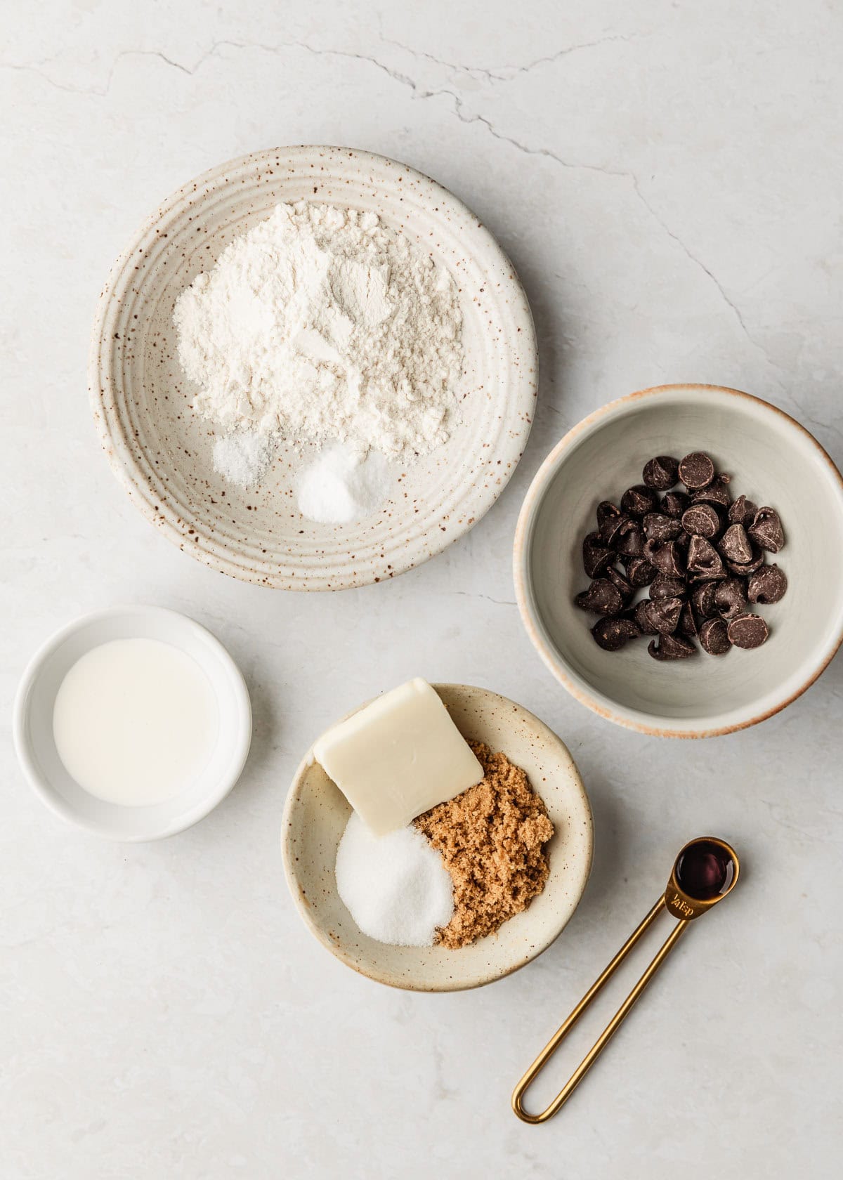 White bowls of flour, chocolate chips, heavy cream, brown sugar, white sugar, and butter on a white counter next to a gold teaspoon of vanilla extract.