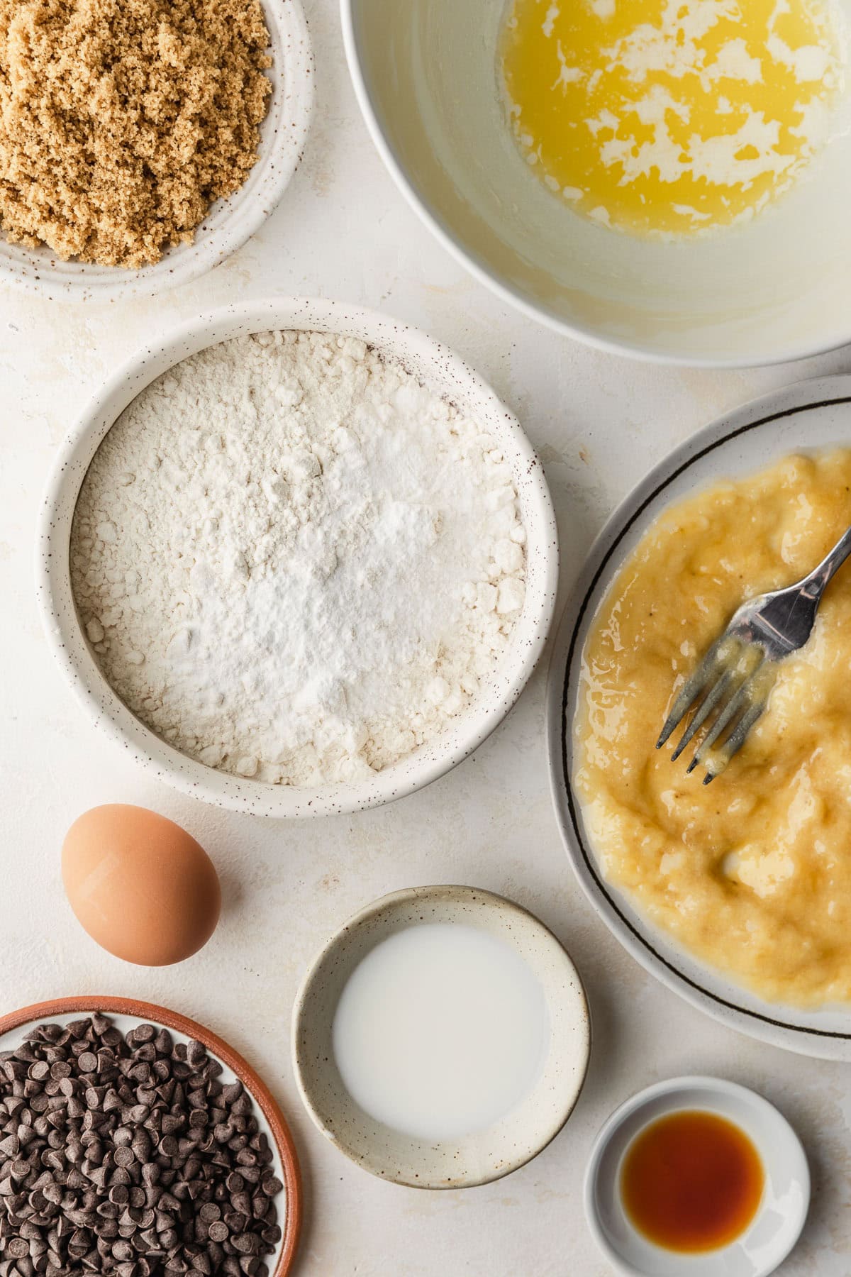 White bowls of melted butter, mashed bananas, flour, brown sugar, milk, vanilla, chocolate chunks, and an egg on a white counter.