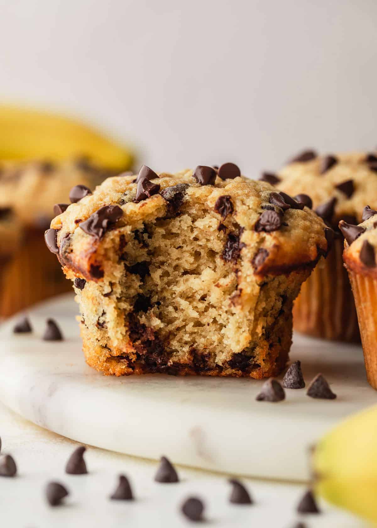 A muffin with a bite taken out of it on a white platter next to rows of small batch chocolate chip banana muffins and bananas with a light beige background.