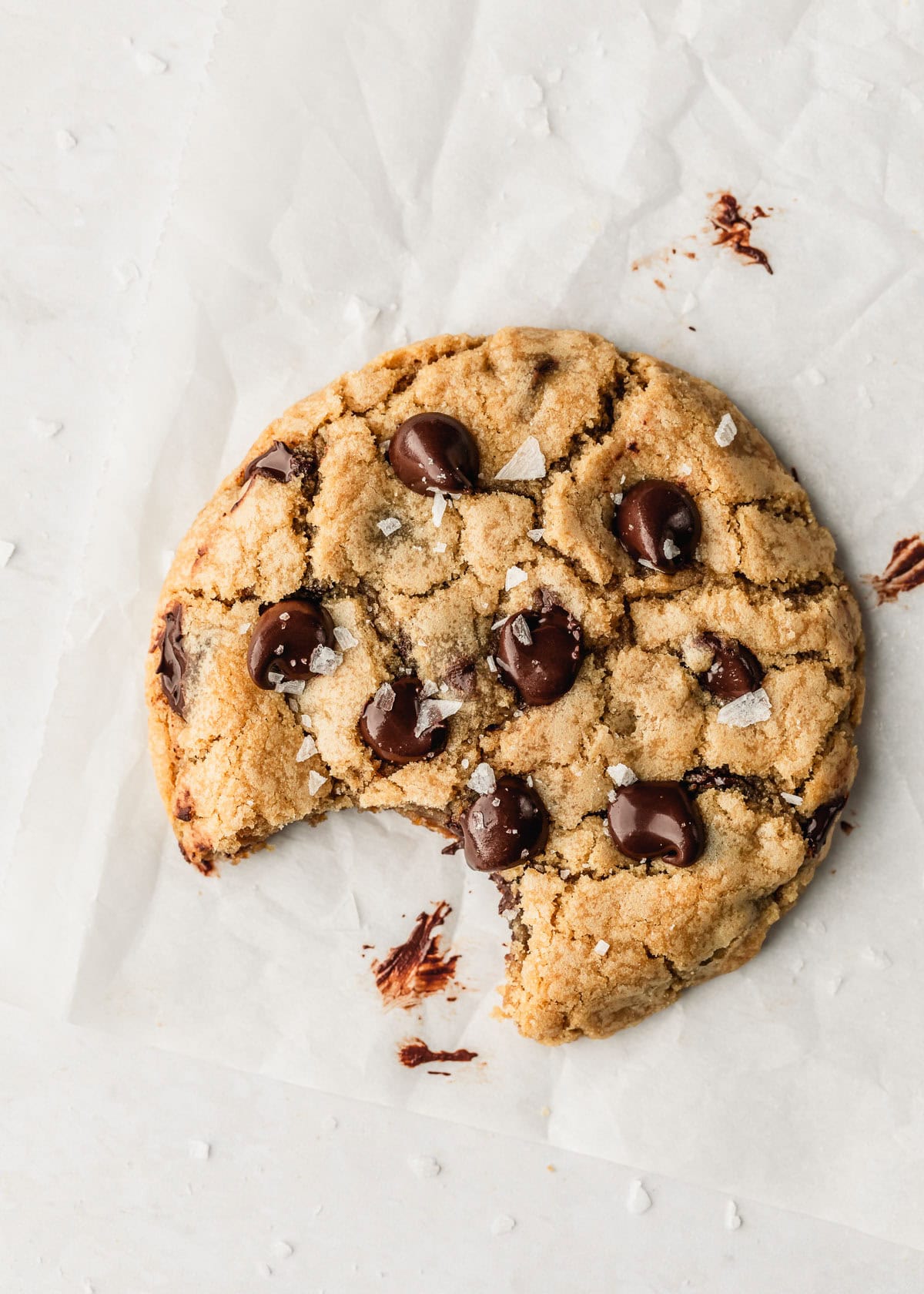 One chocolate chip cookie with a bite taken out of it on a piece of parchment paper with a white marble background.