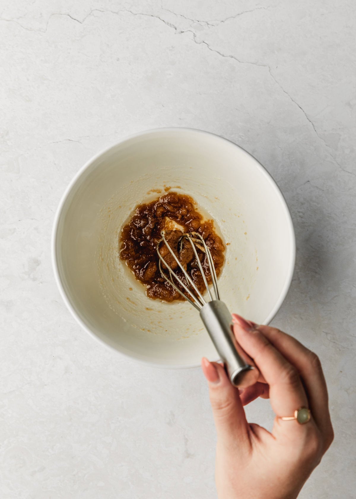 A hand using a whisk to mix butter and sugars in a white bowl.