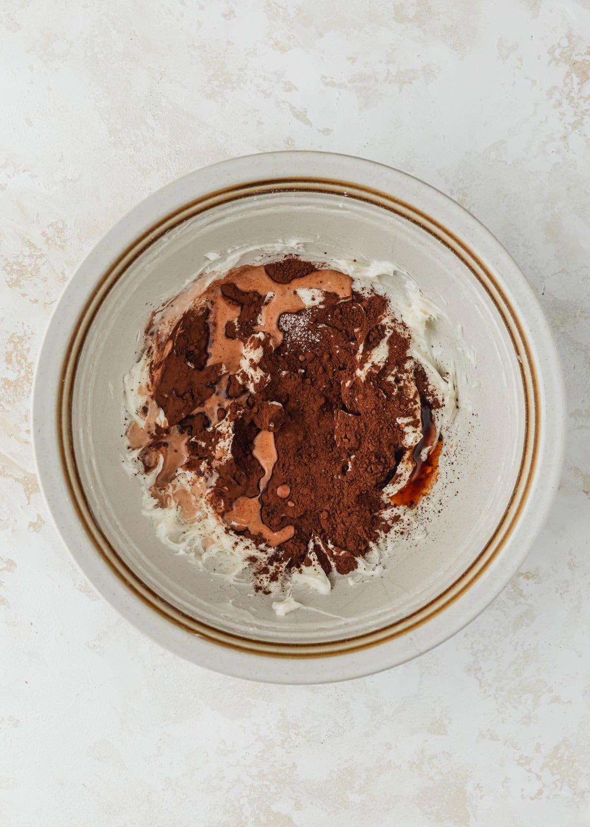 A white bowl of creamed butter and sugar, cocoa, milk, and vanilla on a beige table.