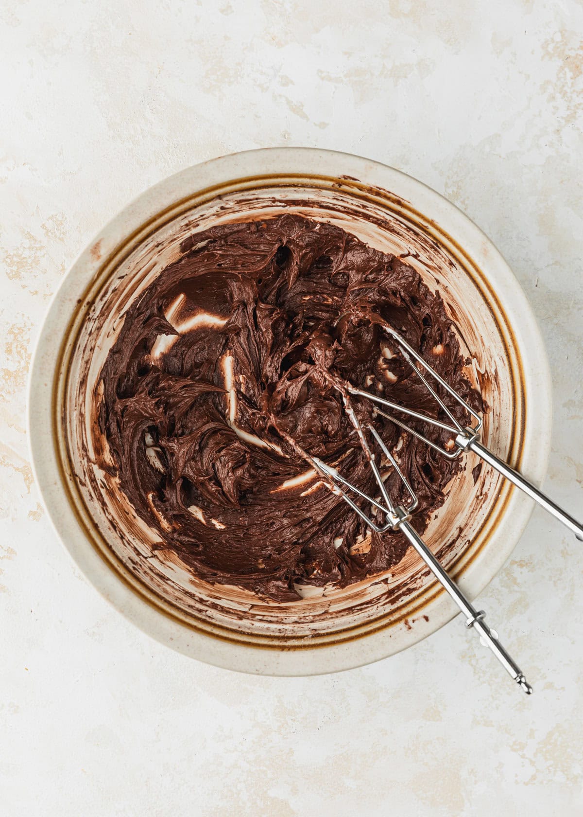 A white bowl of small batch chocolate buttercream frosting and beaters on a tan counter.