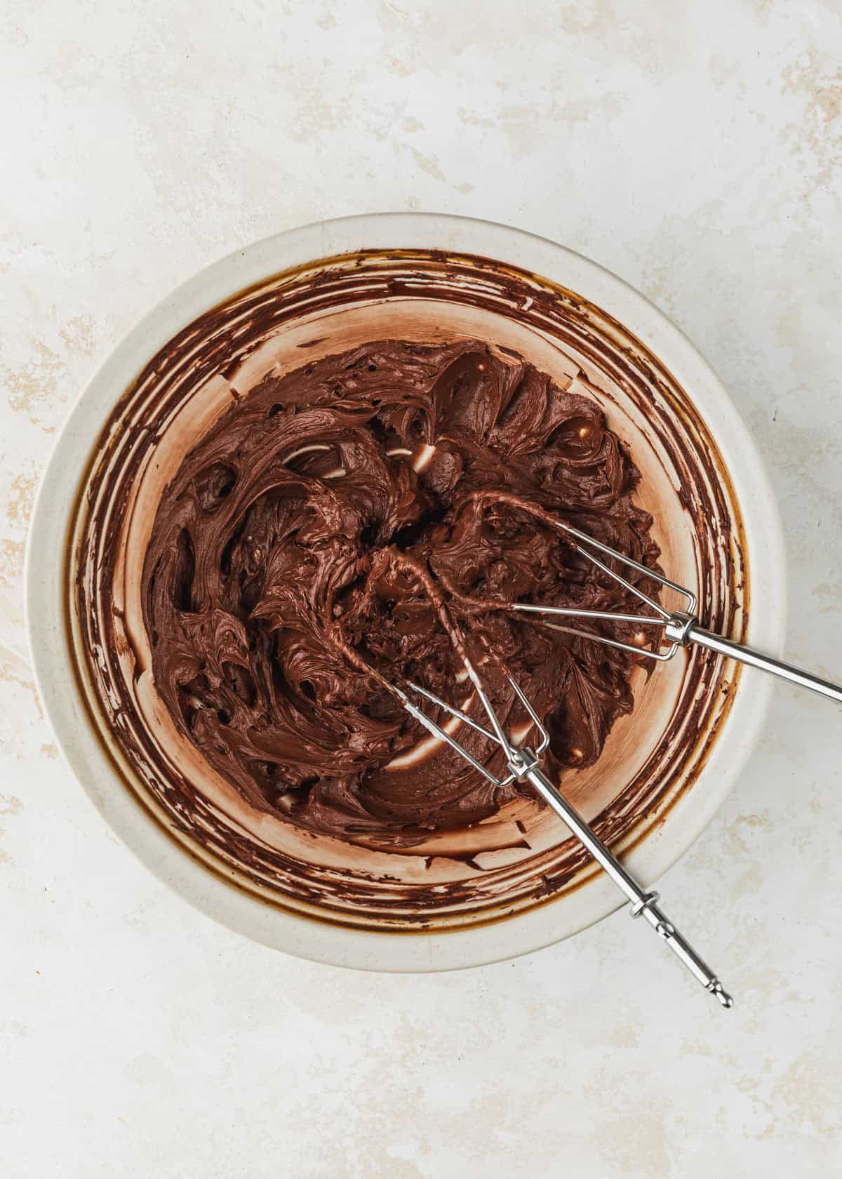 A white bowl of whipped small batch chocolate buttercream frosting and beaters on a beige counter.